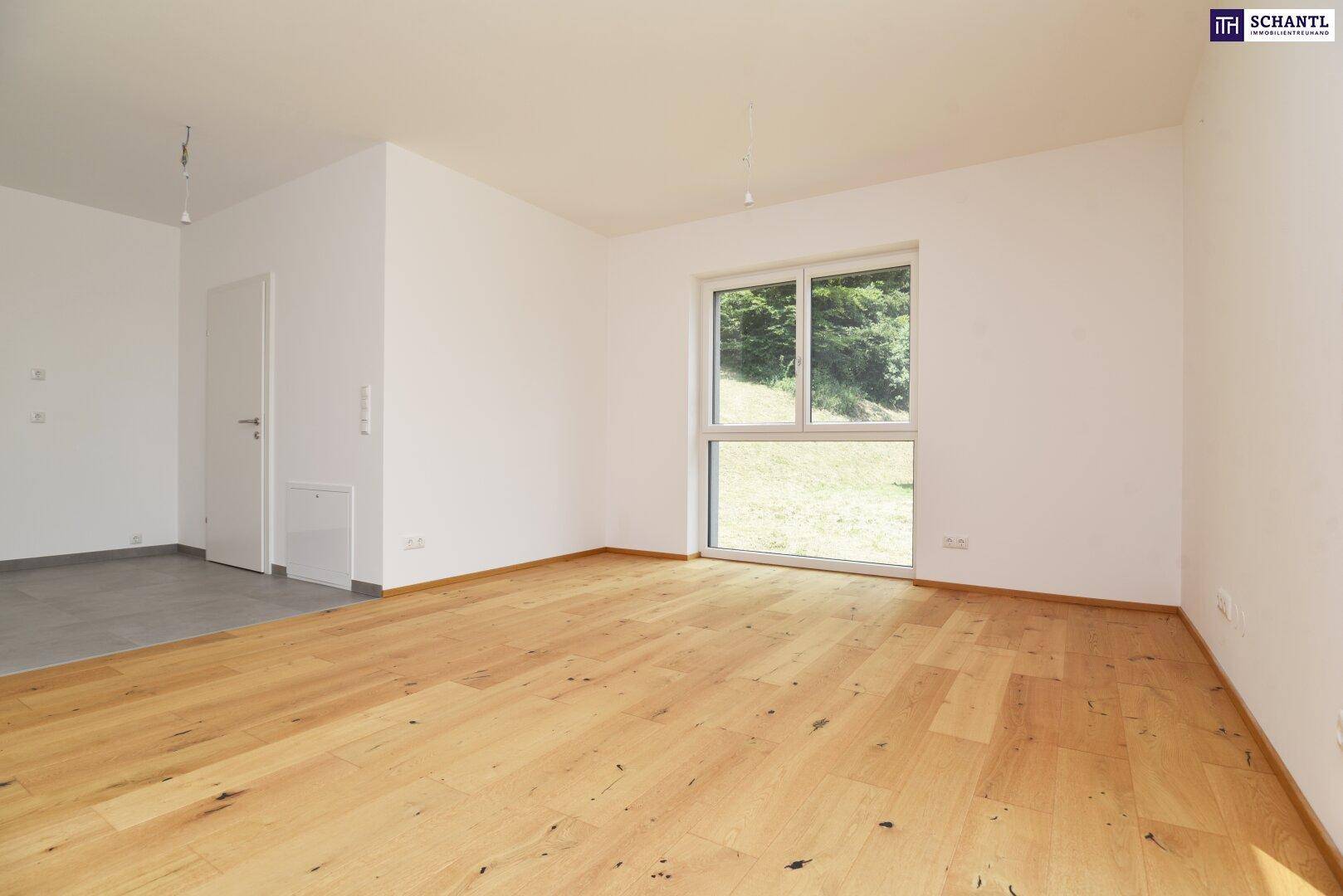 Geräumiges Wohnzimmer mit hellem Holzboden und großem Fenster mit Blick ins Grüne.