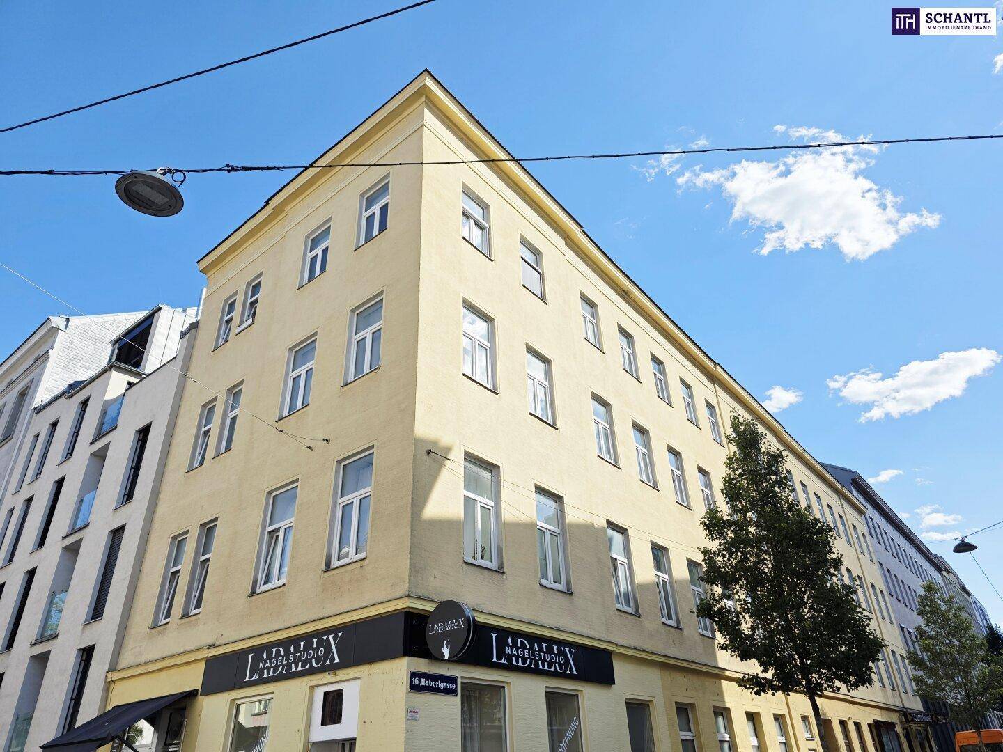 Eckgebäude mit gelber Fassade, vielen Fenstern und Geschäften im Erdgeschoss unter klarem Himmel.