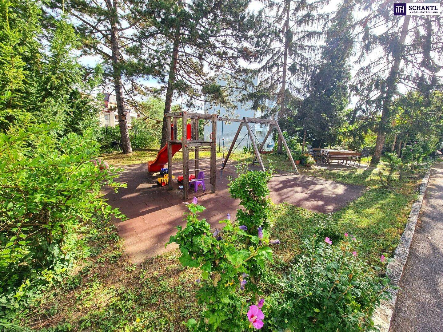 Grüner Gemeinschaftsgarten mit Spielplatz, Rutsche und Schaukeln unter hohen Bäumen.