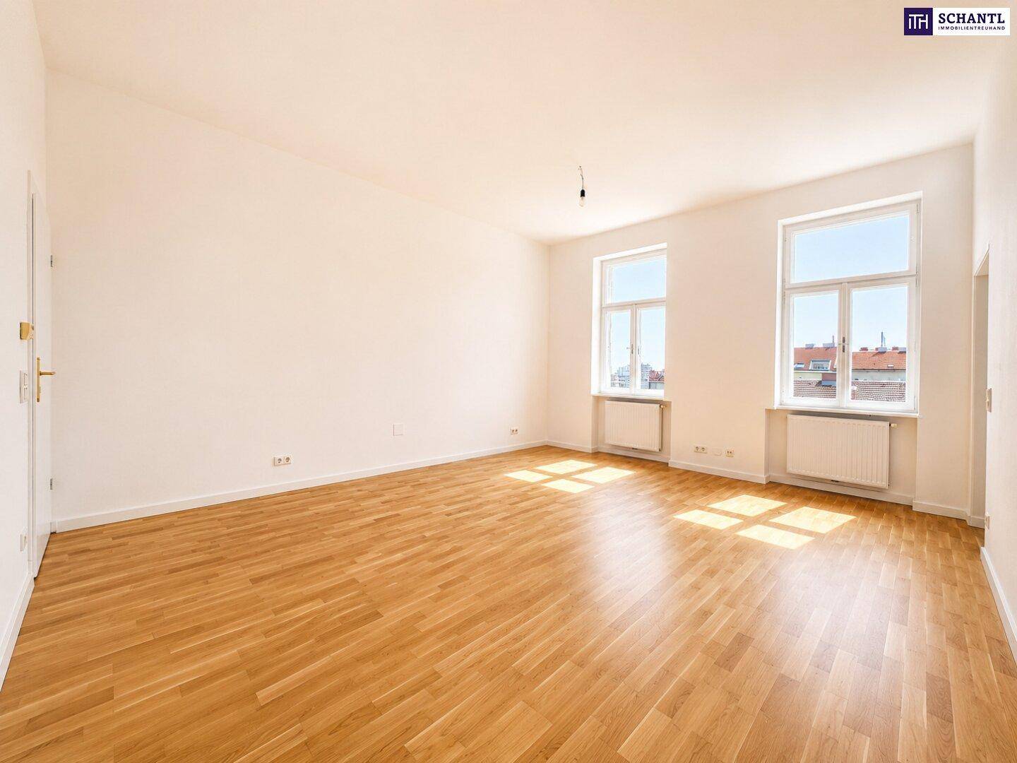 Lichtdurchfluteter Raum mit klassischem Parkettboden und Blick auf die umliegenden Gebäude durch zwei Fenster.