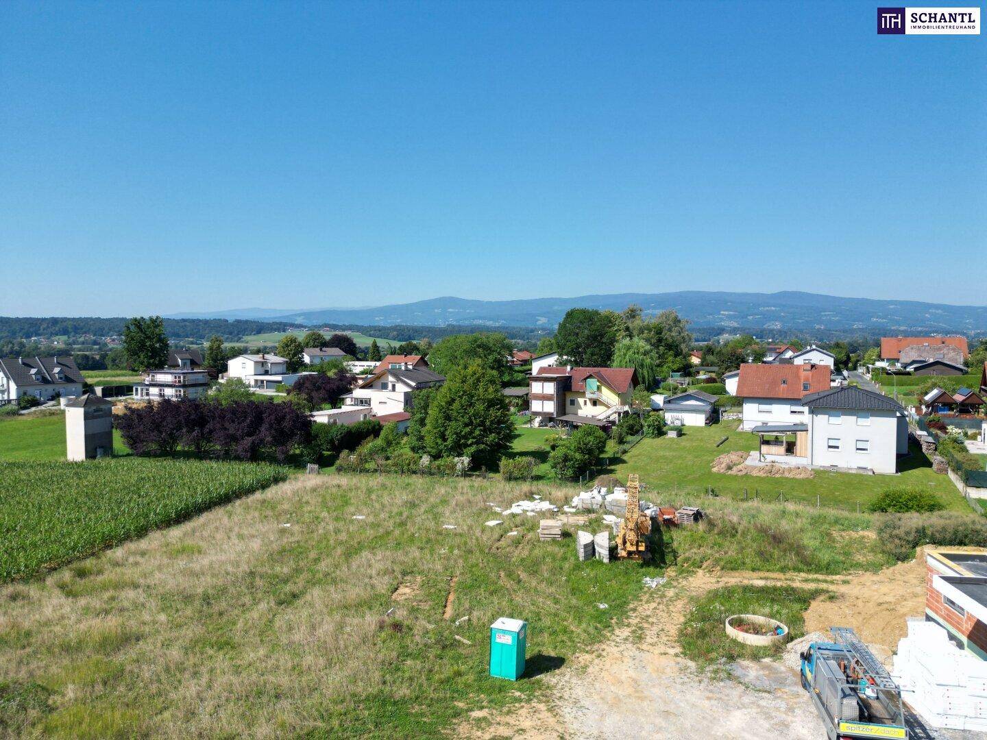 Panoramablick aus der Luft auf eine Baustelle, umgeben von Wohngebäuden und weiten Feldern mit Bergen im Hintergrund.