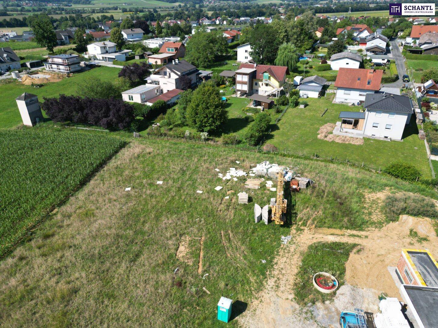 Luftaufnahme einer Baustelle mit Baumaterialien und einem Kran, umgeben von Wohnsiedlungen und landwirtschaftlichen Flächen.
