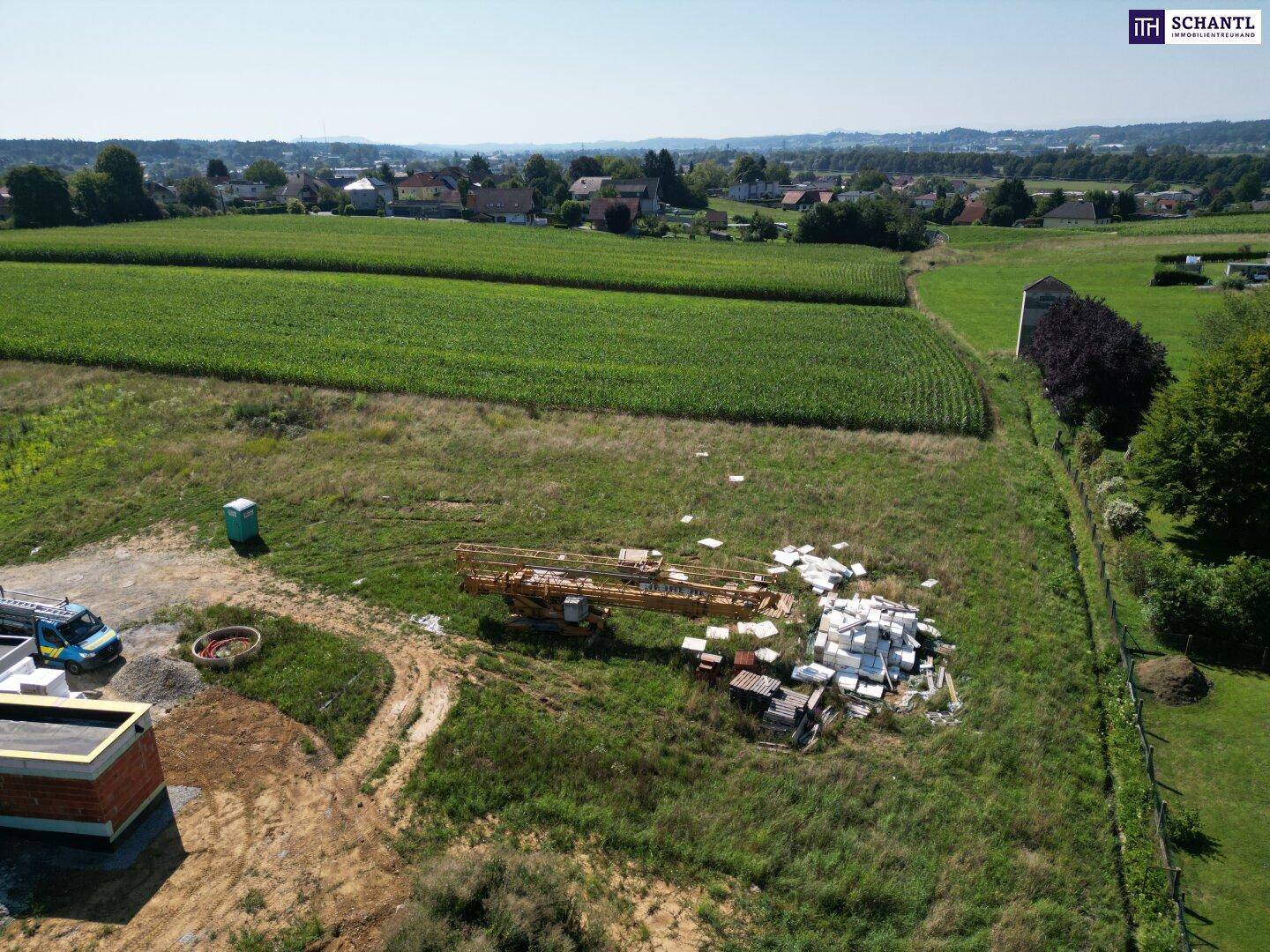 Luftaufnahme einer Baustelle neben einem Maisfeld, umgeben von weiteren Feldern und einer ländlichen Wohnsiedlung.