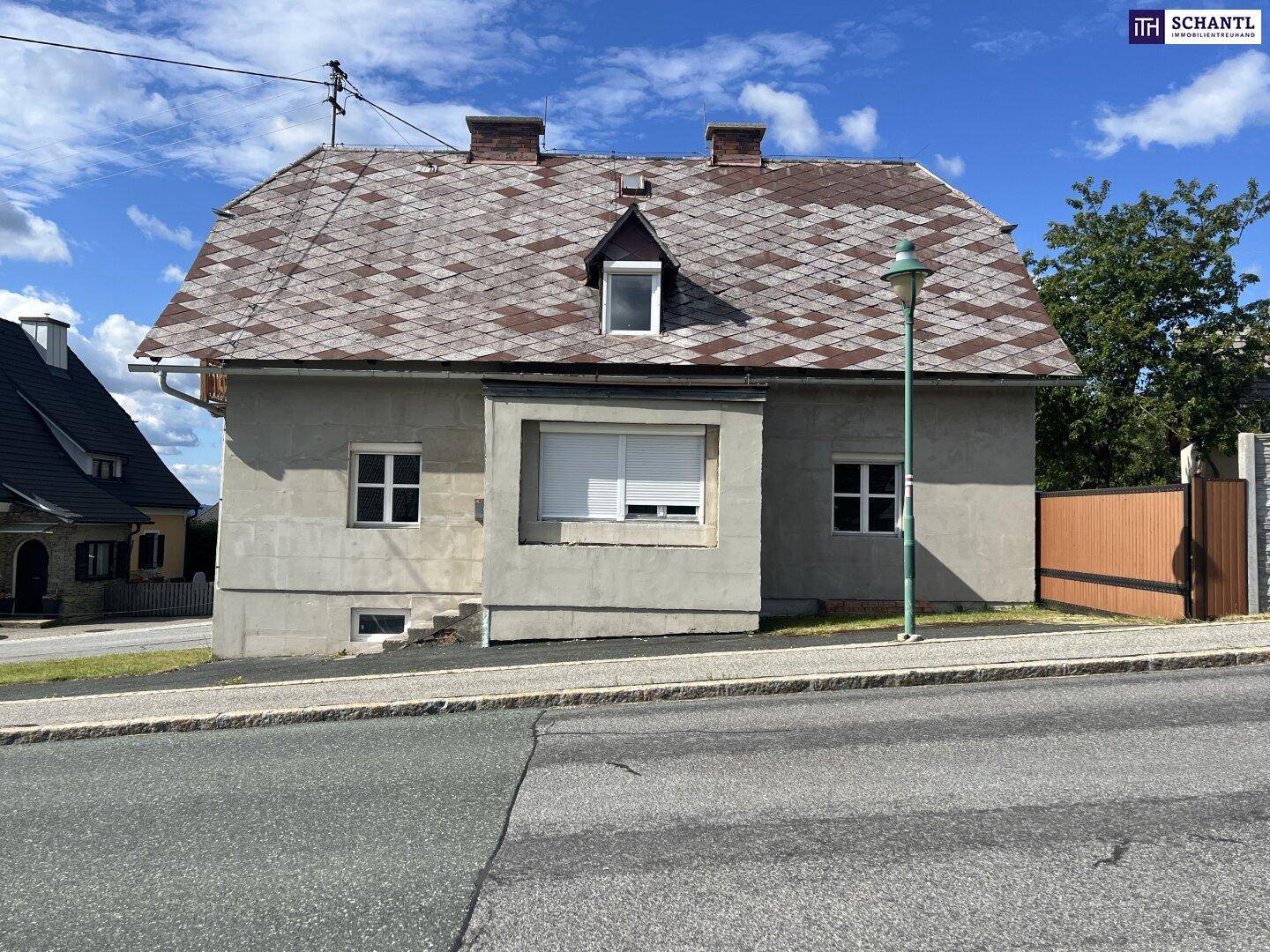 Älteres Haus mit Satteldach und Schindeln, direkt an einer Straße gelegen, unter bewölktem Himmel.