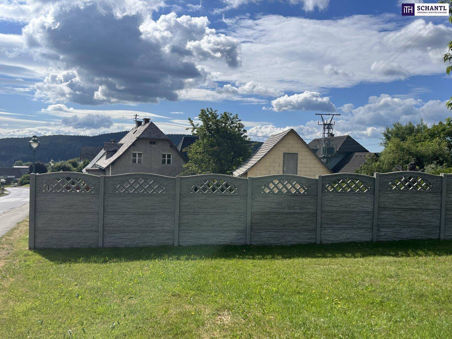 Robuster Betonzaun begrenzt ein grünes Grundstück mit Blick auf Häuser und Hügel im Hintergrund.