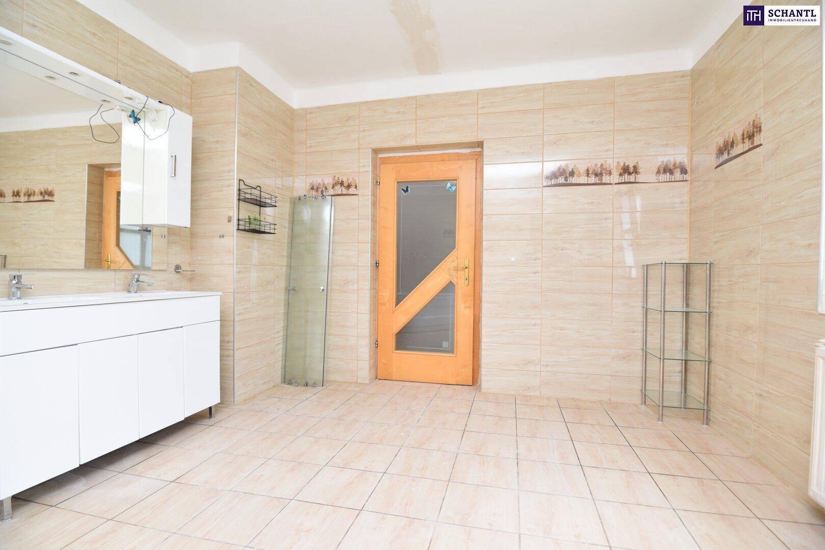Großes Badezimmer mit beigefarbenen Fliesen, Doppelwaschbecken und Holztür, renovierungsbedürftig.