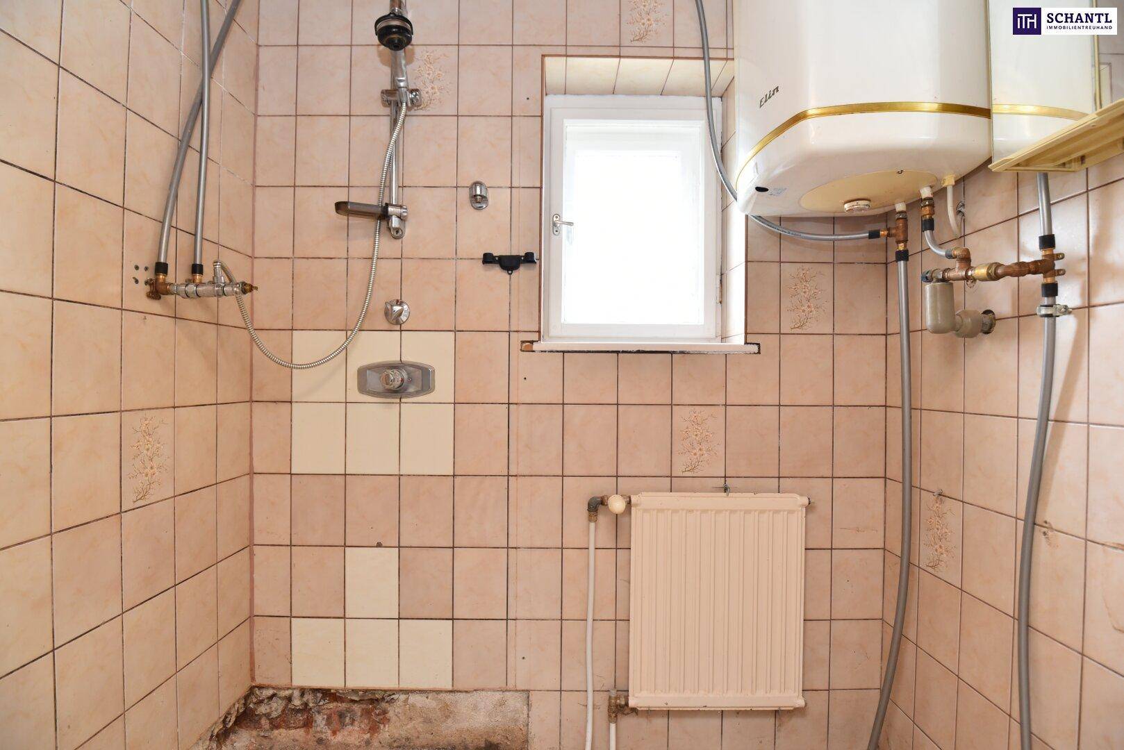 Badezimmer mit Duschbereich, Boiler, Heizkörper und Fenster, das Renovierungsbedarf aufweist.