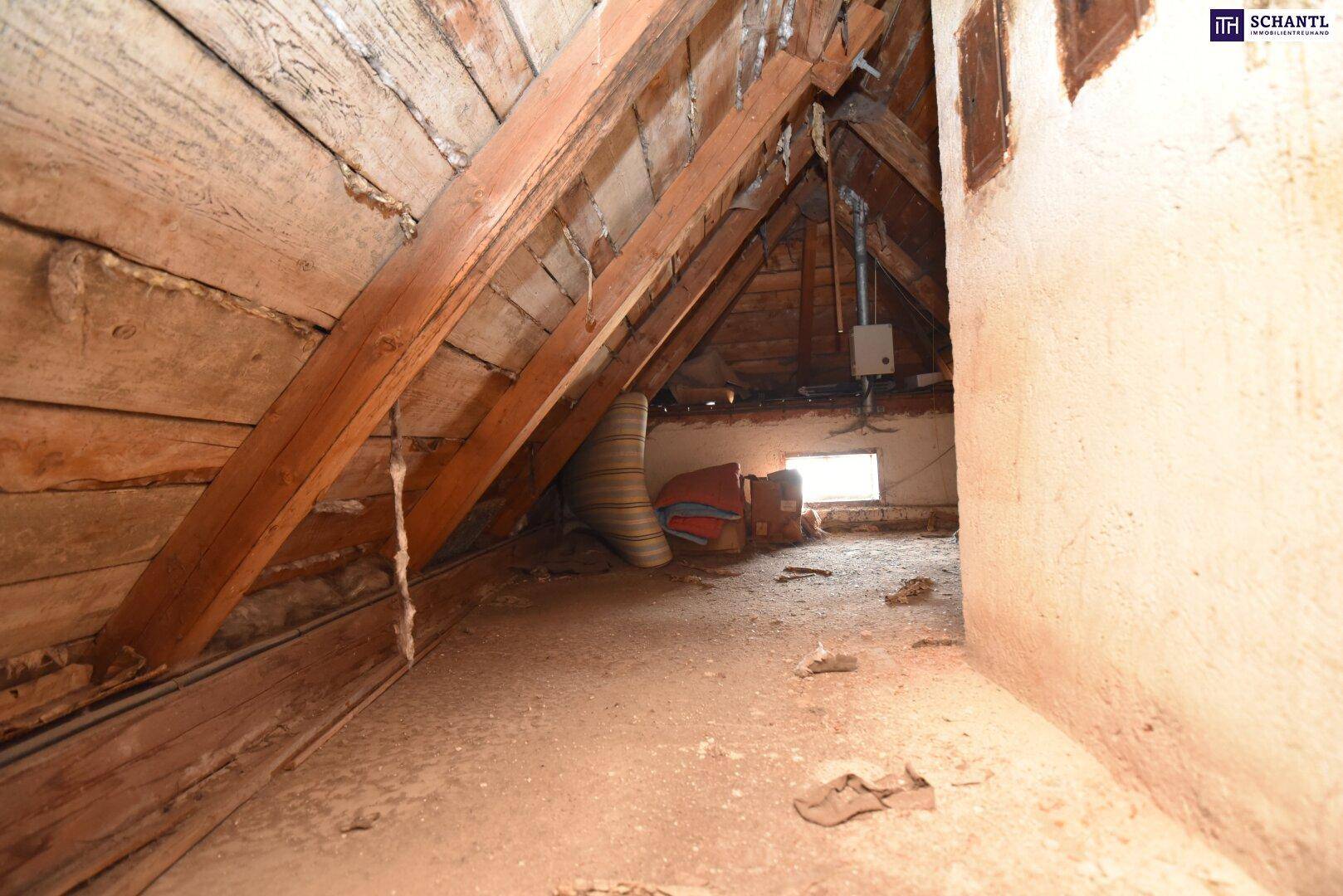 Unausgebauter Dachboden mit sichtbaren Holzbalken, einem kleinen Fenster und gelagerten Gegenständen.