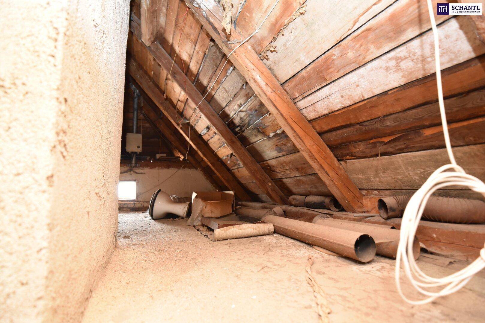 Unausgebauter Dachboden mit sichtbaren Holzbalken, Rohrleitungen und einem kleinen Fenster.
