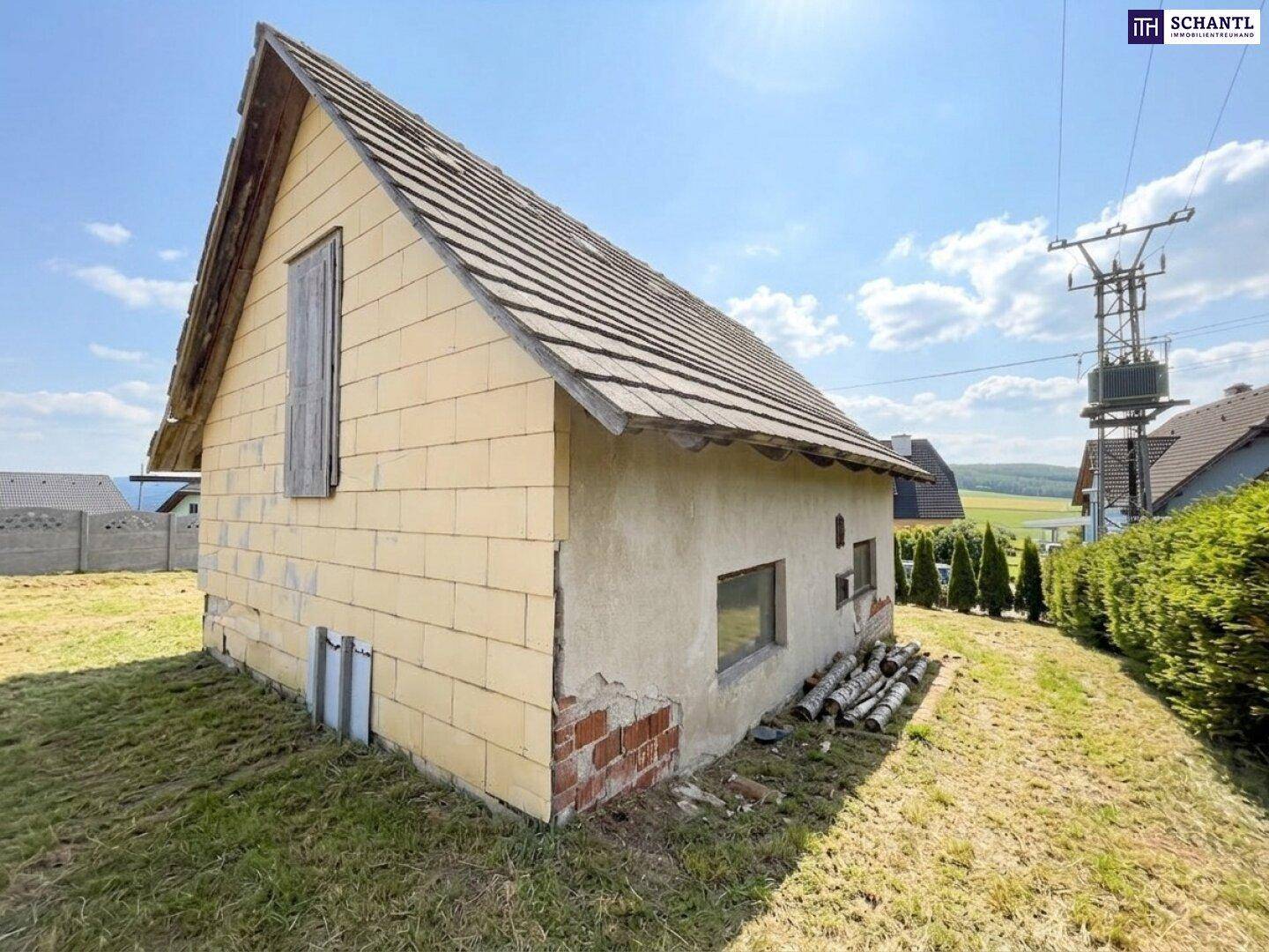 Kleines Nebengebäude mit Satteldach und verputzten Wänden, umgeben von trockenem Gras und Bäumen.