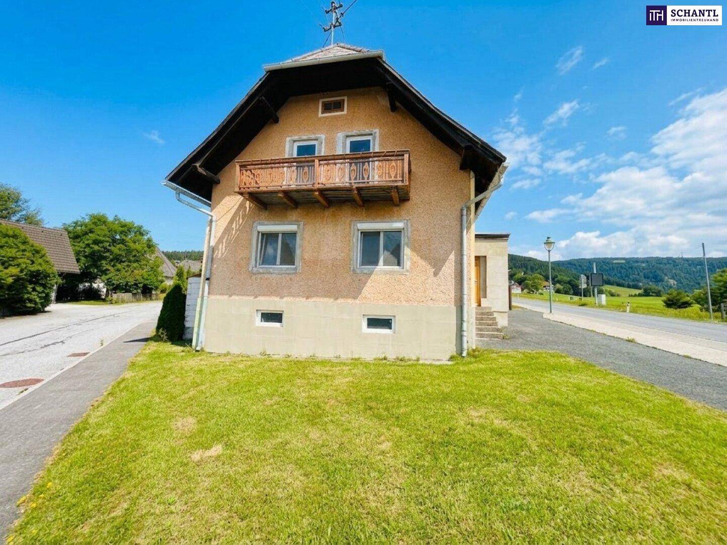 Traditionelles Haus mit Satteldach und kleinem Balkon, umgeben von Grünflächen unter blauem Himmel.