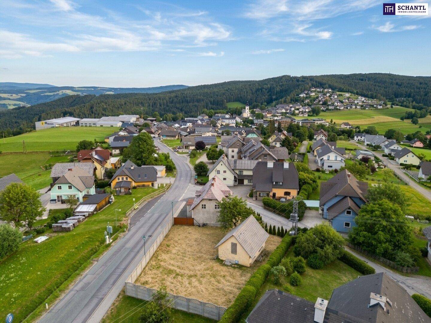 Weitläufige Luftaufnahme eines Dorfes mit vielen Häusern und Feldern, umgeben von Wäldern und Bergen.
