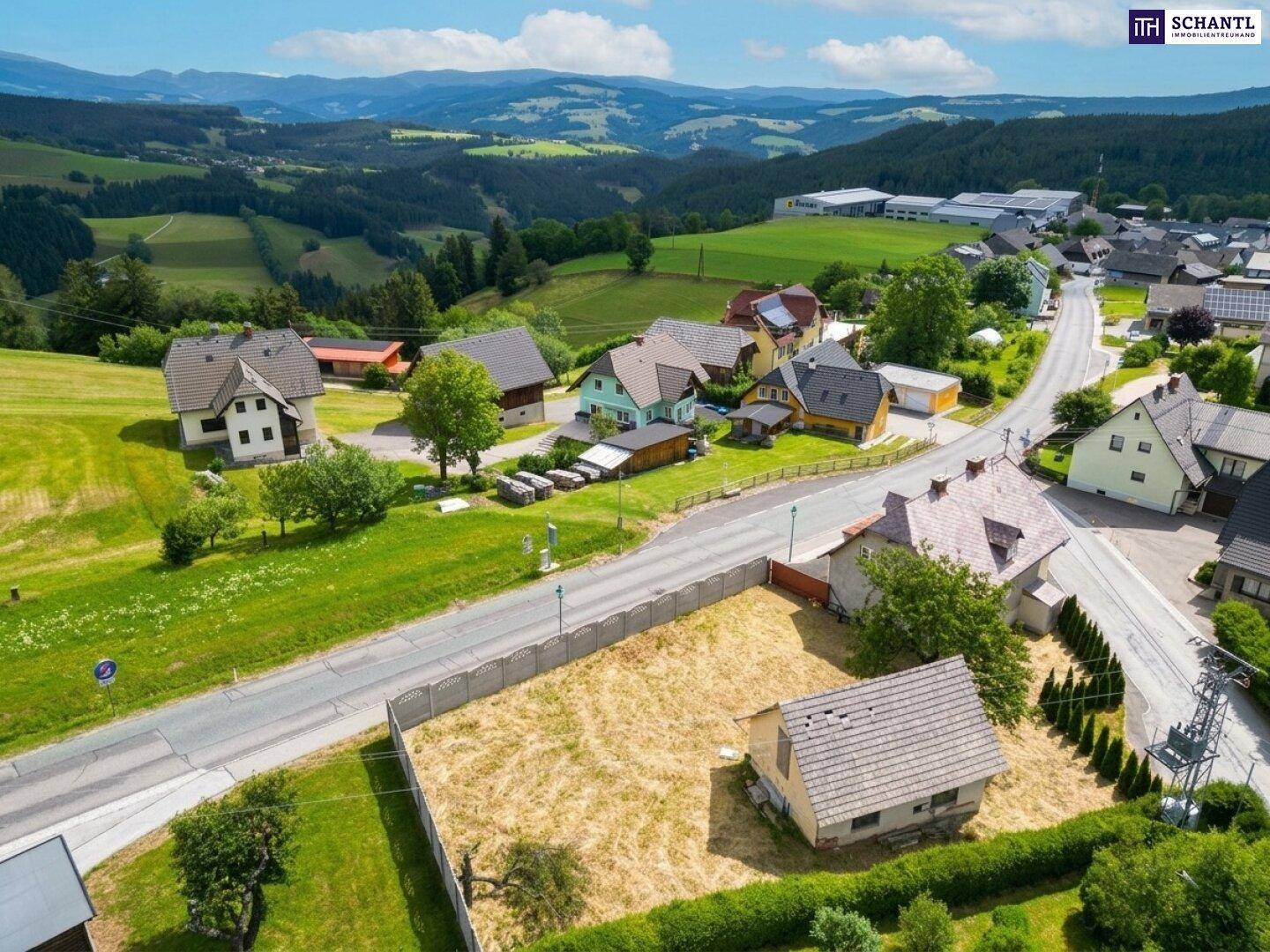 Vogelperspektive auf ein ländliches Dorf mit Häusern, Straßen und viel Grün, umgeben von Wäldern.