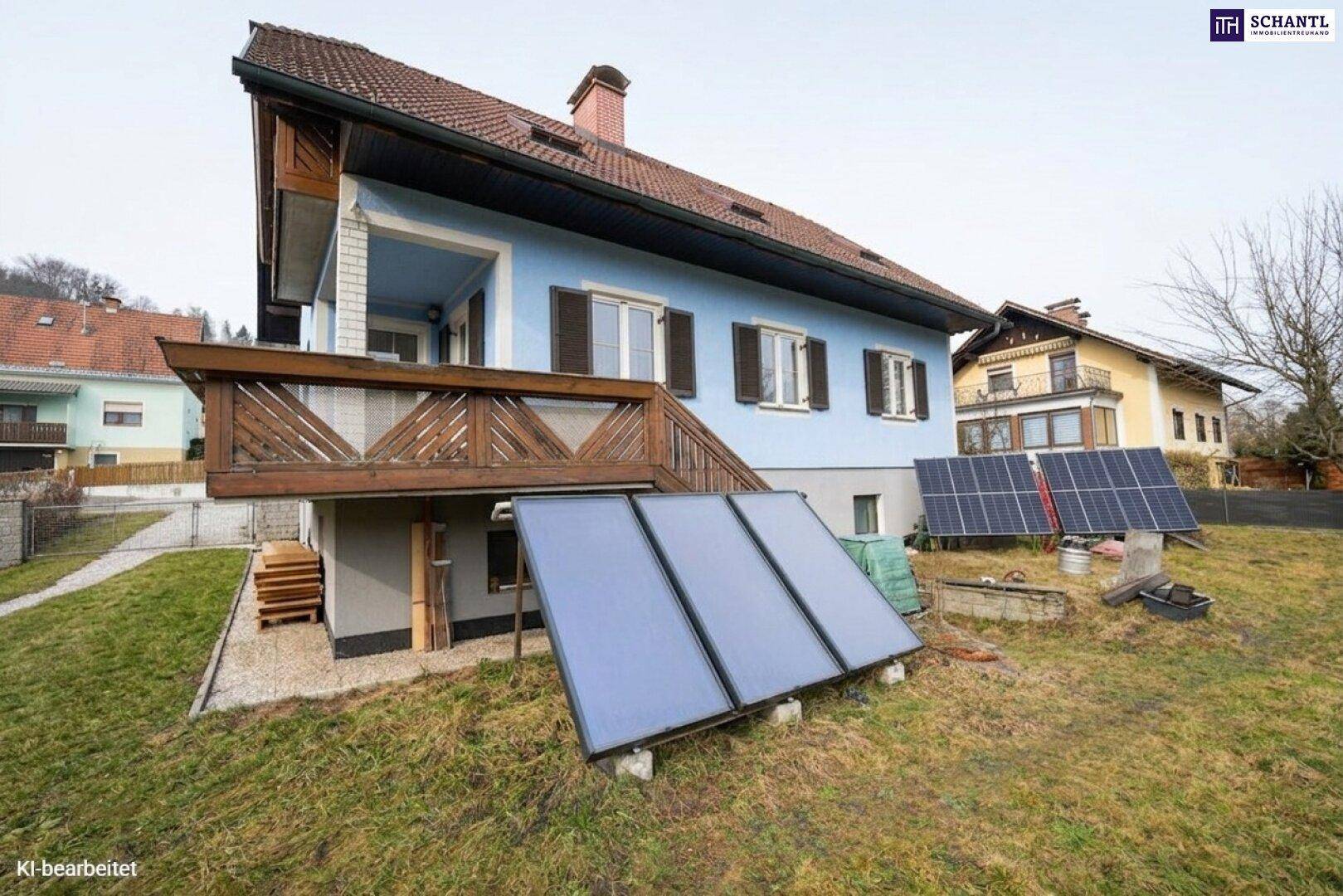 Rückseite des Hauses mit Balkon und Solaranlagen auf dem Rasen, umgeben von grüner Landschaft.