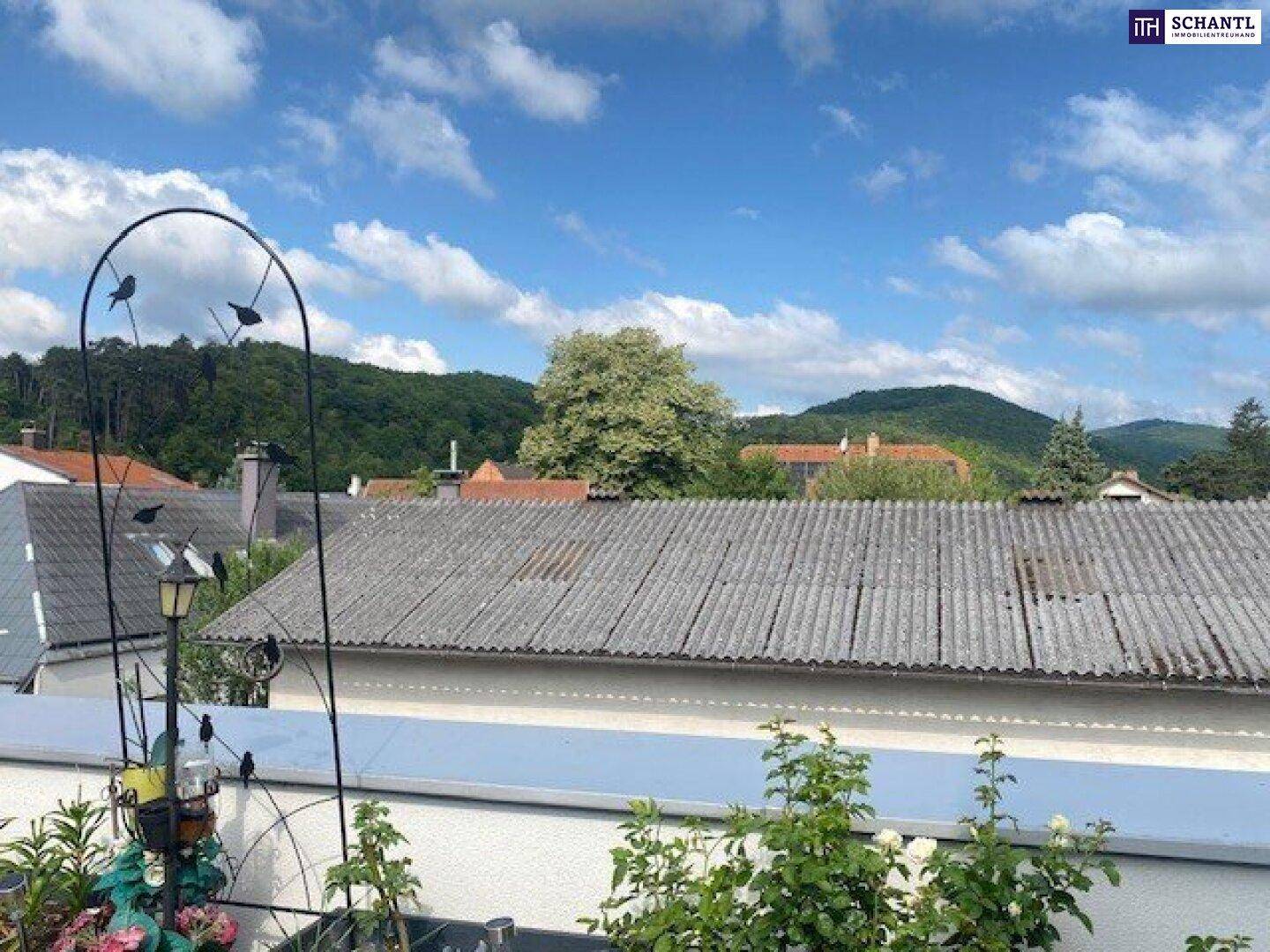 Blick von einem Balkon über Dächer und grüne Hügel unter blauem Himmel.