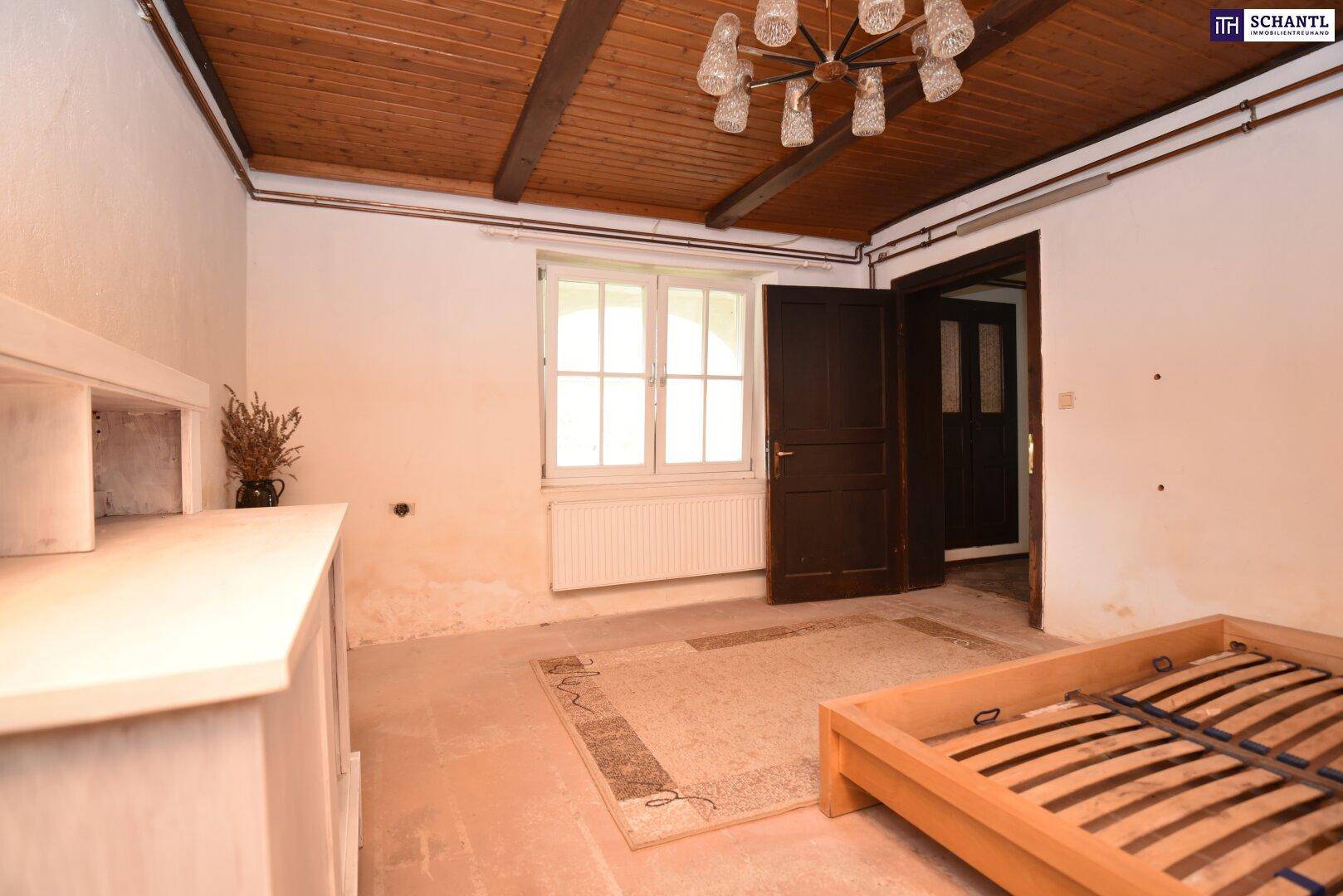Schlafzimmer mit Holzbalkendecke, Fenster und einer Tür, die zu einem weiteren Raum führt.
