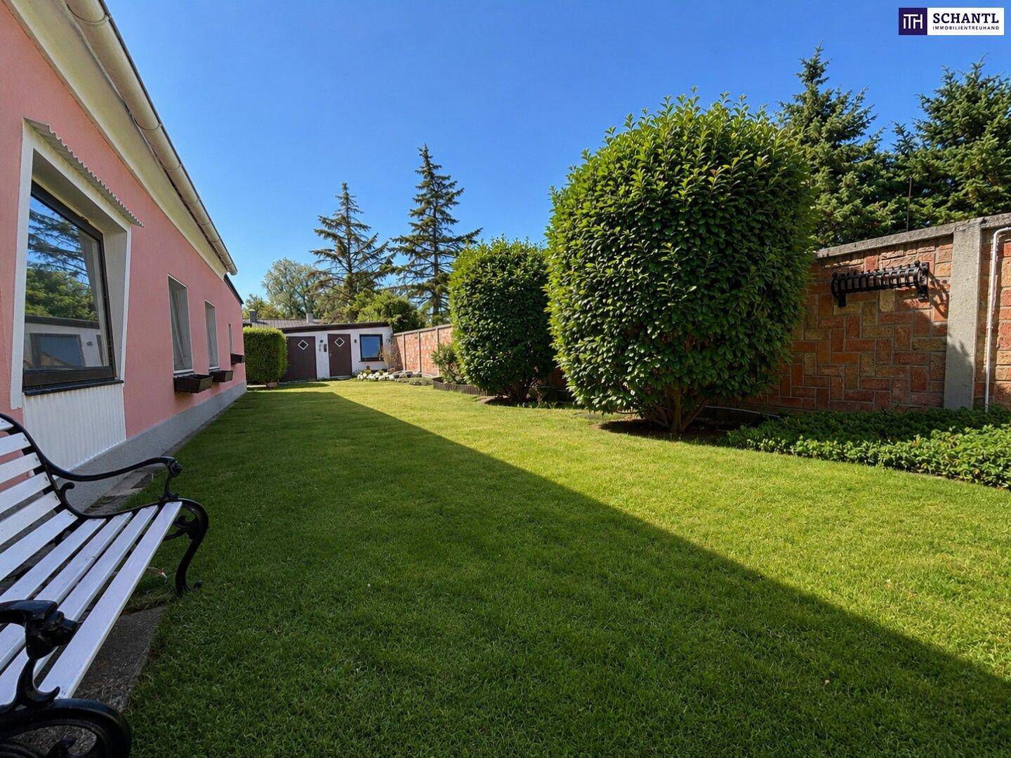 Sonniger Gartenbereich mit Rasen, Hecken und einer Gartenbank entlang der Hauswand.