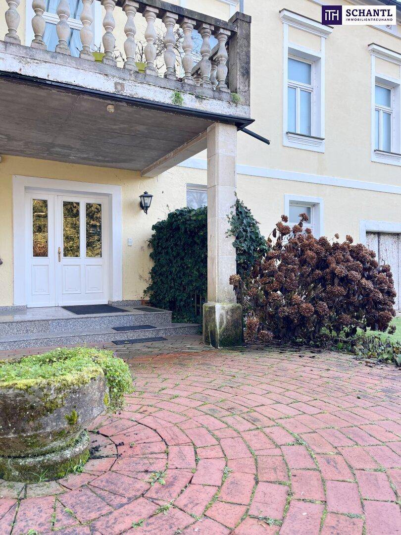 Eingangsbereich des Anwesens mit gepflastertem Vorplatz, historischem Balkon und einem steinernen Pflanzgefäß.