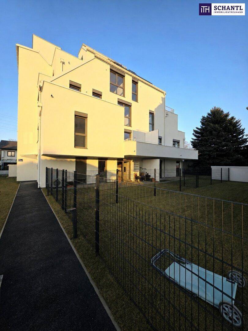 Seitenansicht des modernen Neubaus mit weißer Fassade, umzäuntem Gartenbereich und klarem Himmel.