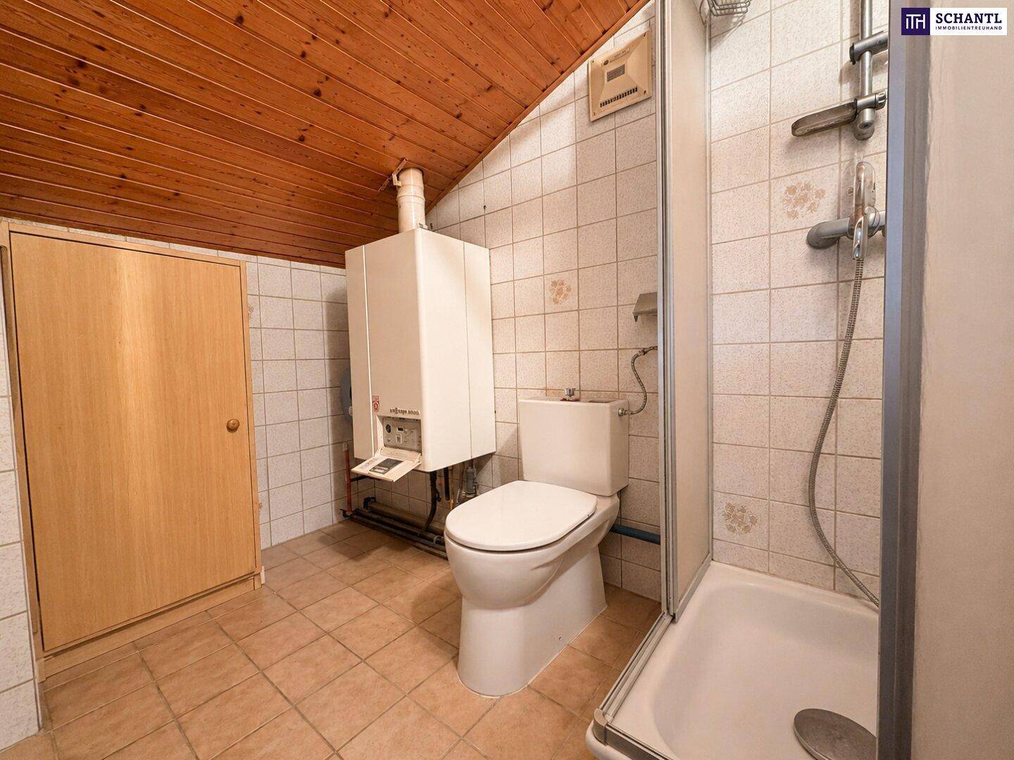 Badezimmer mit Dusche, Toilette und Warmwasserboiler unter einer schrägen Holzdecke.