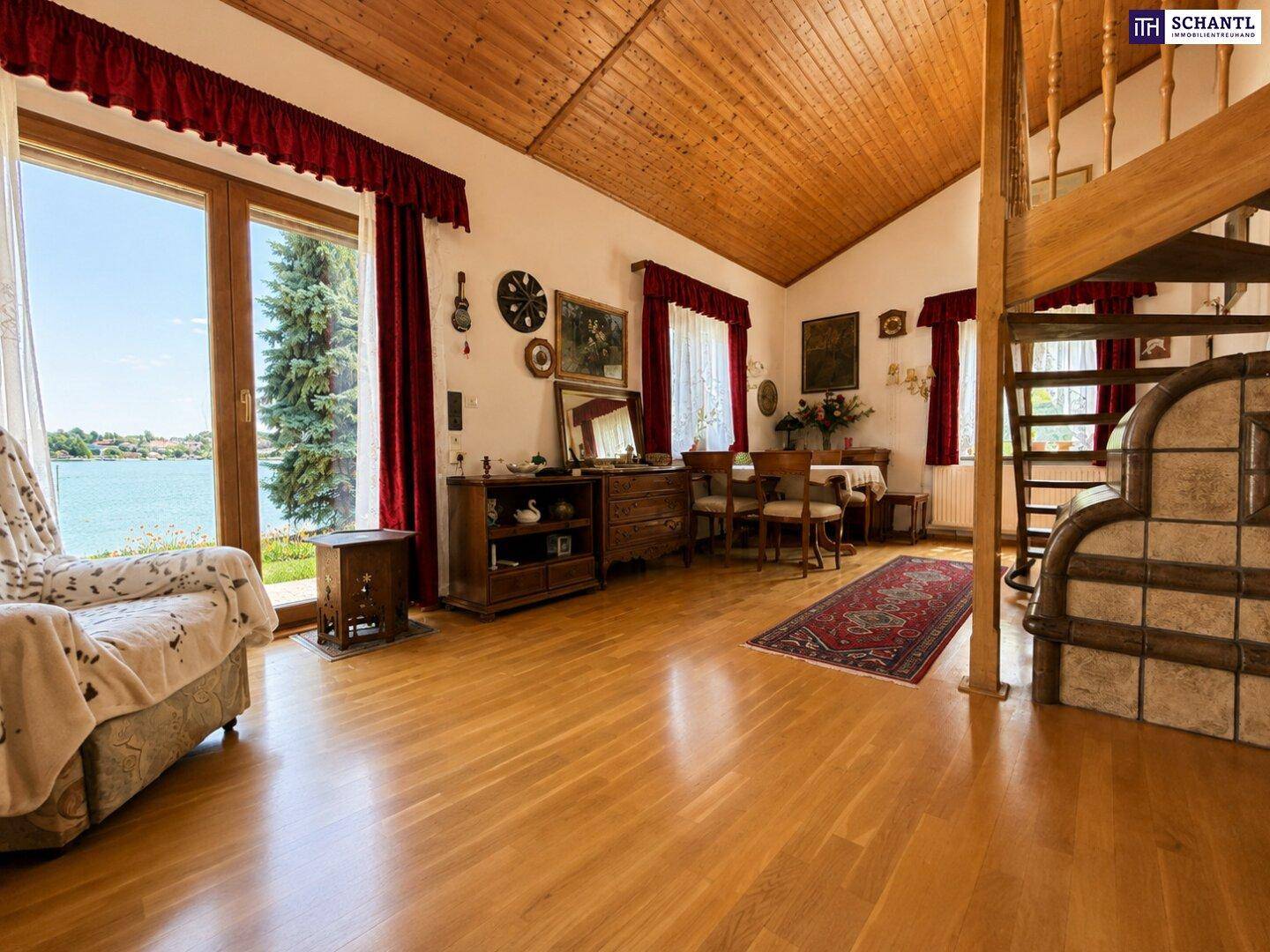 Geräumiges Wohnzimmer mit Holzbalkendecke, Parkettboden und direktem Seeblick durch große Fenster.