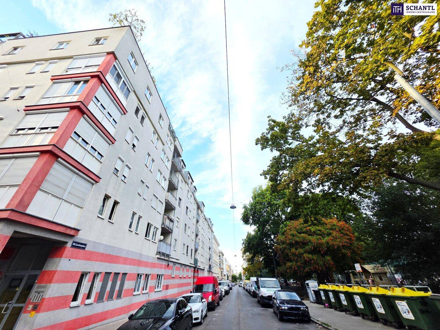 Straßenansicht eines Wohngebäudes mit Bäumen, parkenden Autos und Müllcontainern.