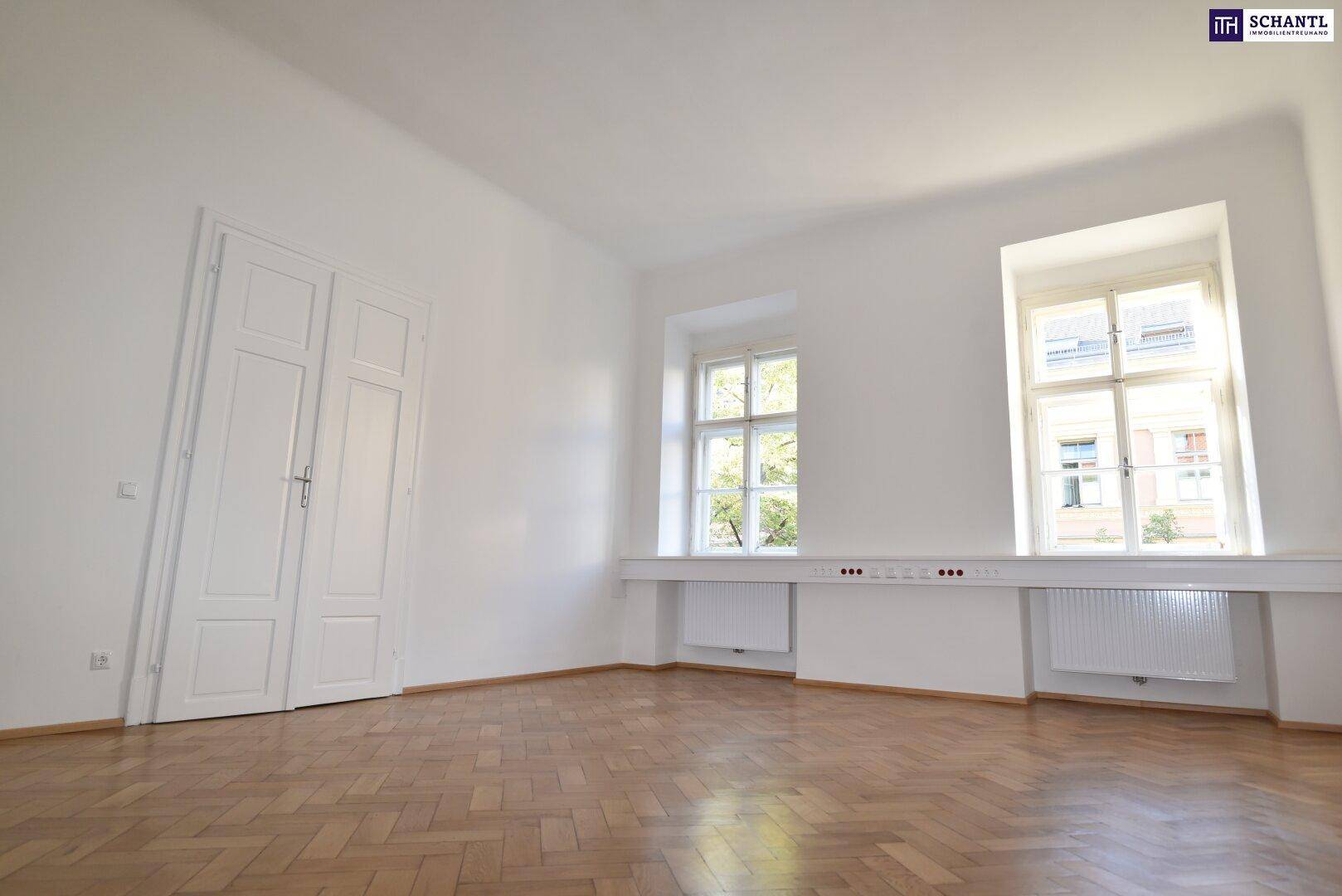 Geräumiger, heller Raum mit Fischgrätparkett, weißen Wänden und zwei Fenstern.