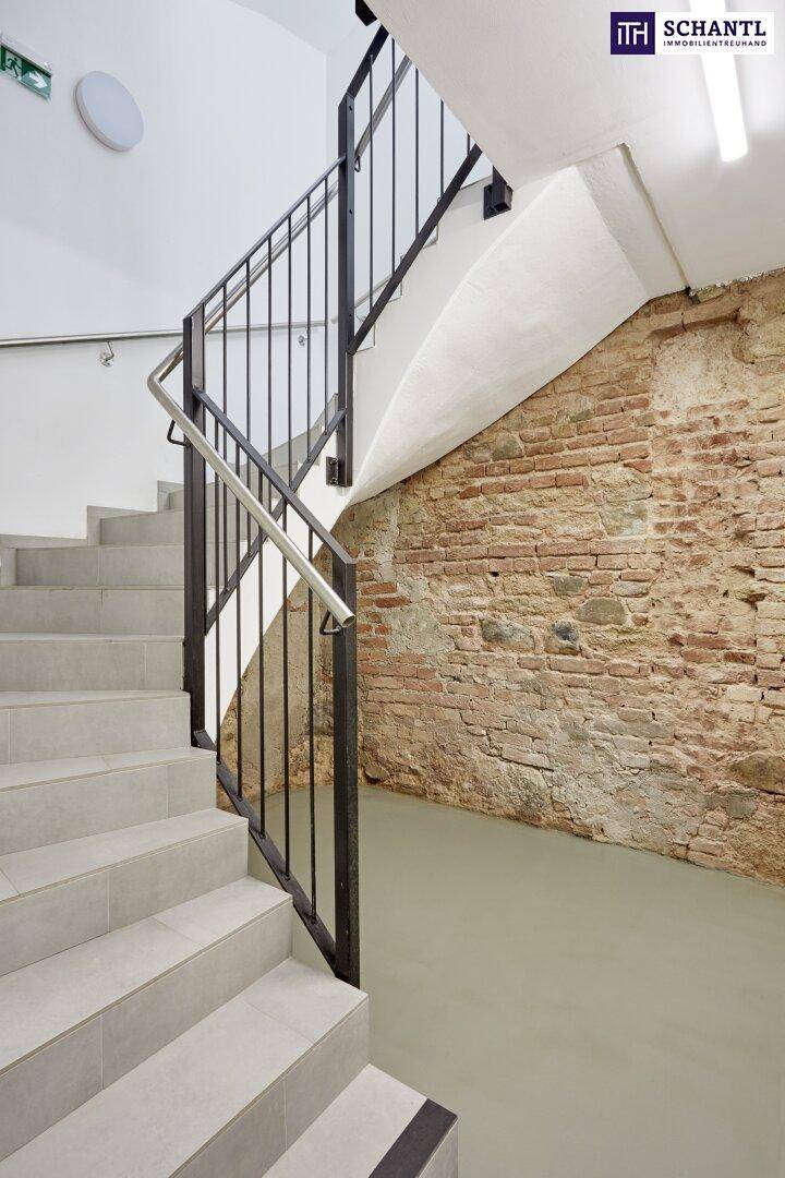 Treppenhaus mit modernen Stufen, Metallgeländer und freigelegter Ziegelwand.