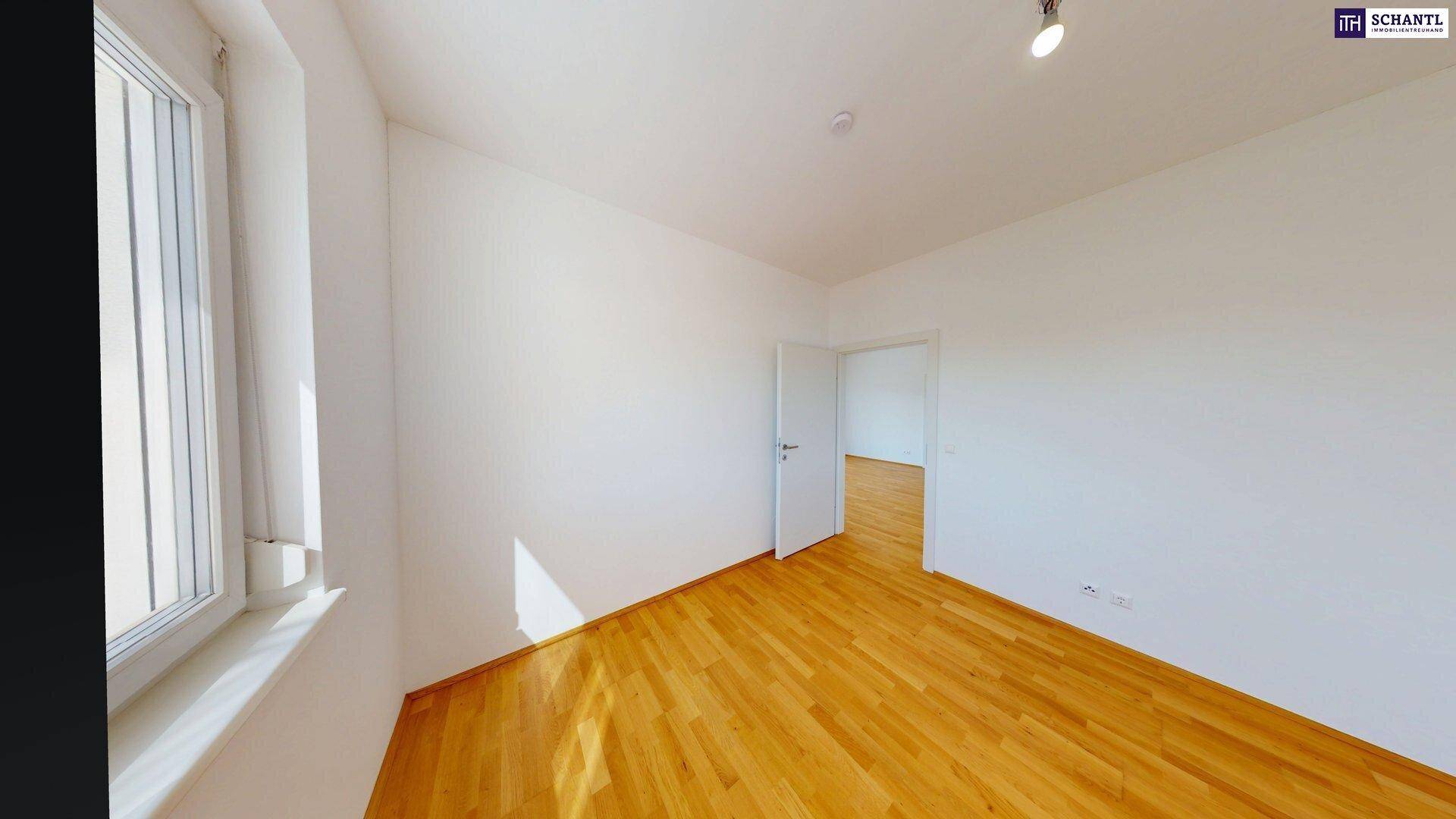 Leerer Raum mit Holzboden und großem Fenster, das viel Tageslicht hereinlässt.