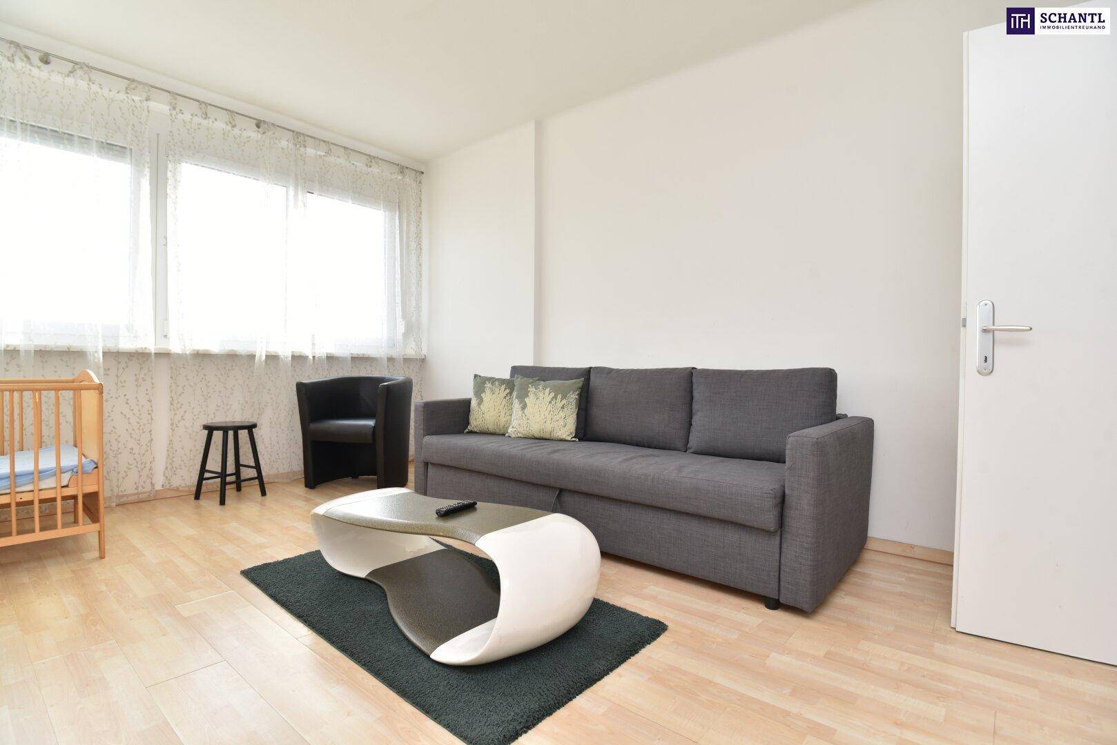 Wohnzimmer mit grauem Sofa, modernem Couchtisch und Babybett am Fenster.