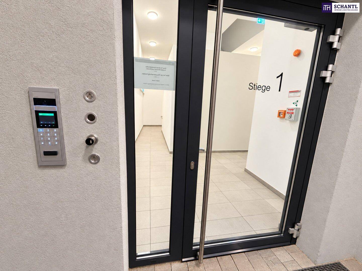 Eingangsbereich des Gebäudes mit Gegensprechanlage und Glastüren, die in den Flur führen.
