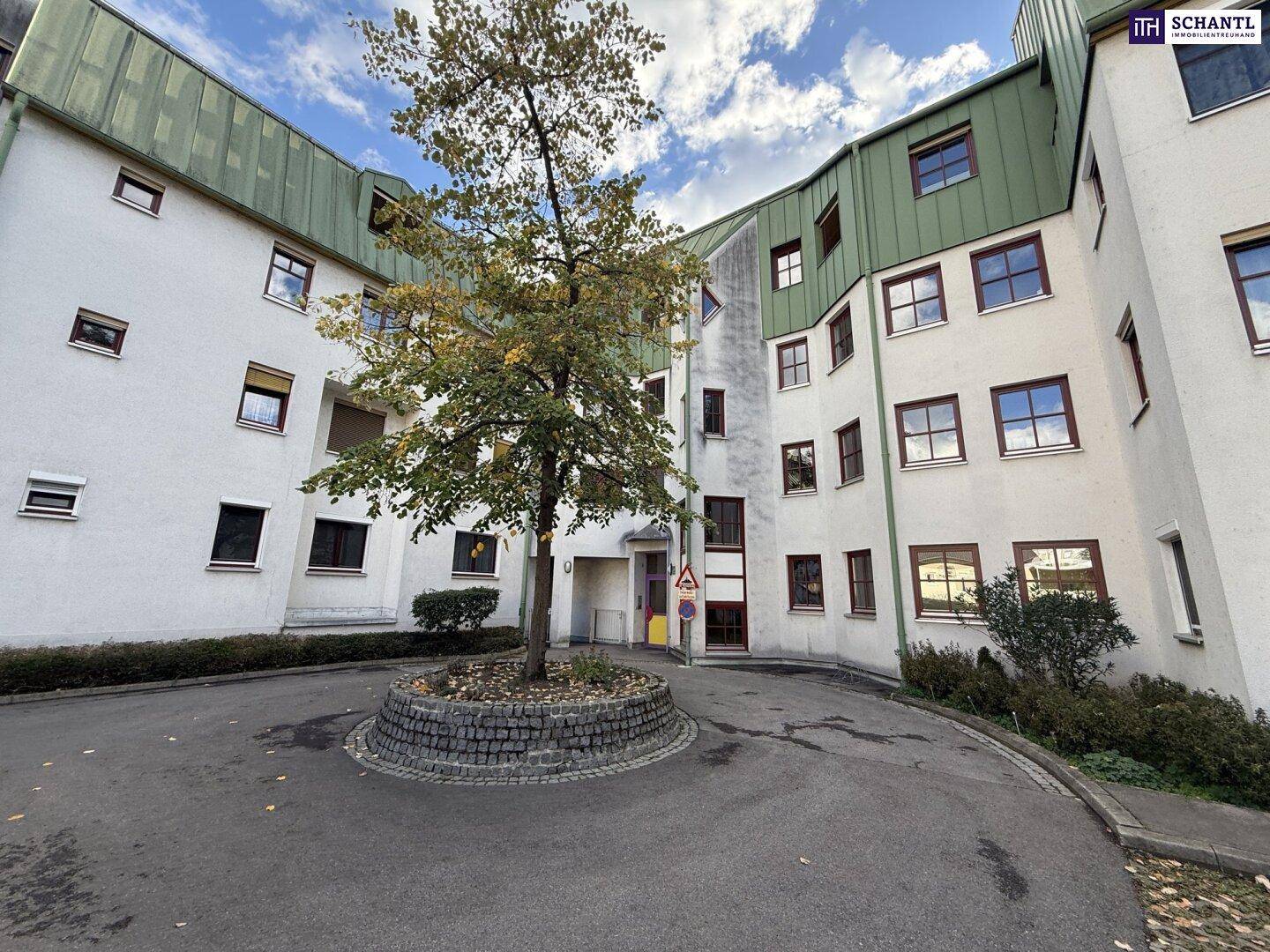 Außenansicht eines Wohngebäudes mit weißer Fassade, grünem Dach und zentralem Baum im Hof.