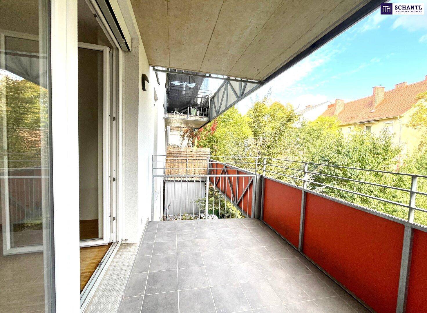 Geräumiger Balkon mit Fliesenboden und roter Brüstung, umgeben von Bäumen und Gebäuden.