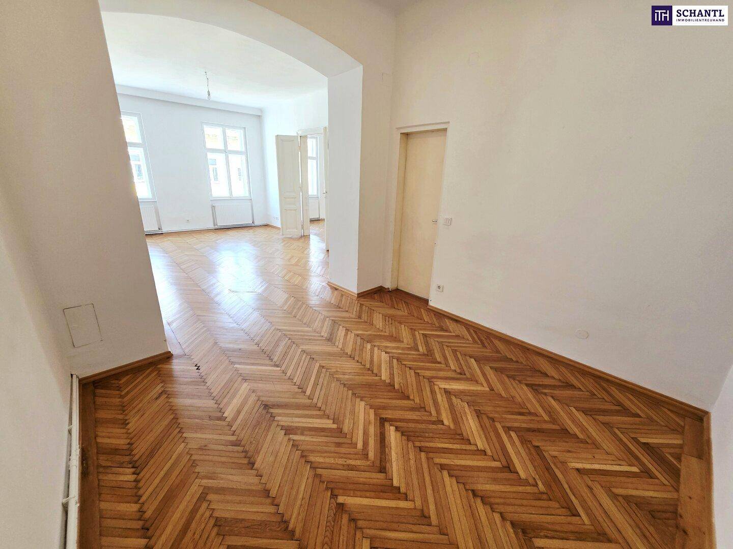 Langer Raum mit Fischgrätparkett und einem offenen Durchgang zu einem weiteren Zimmer.