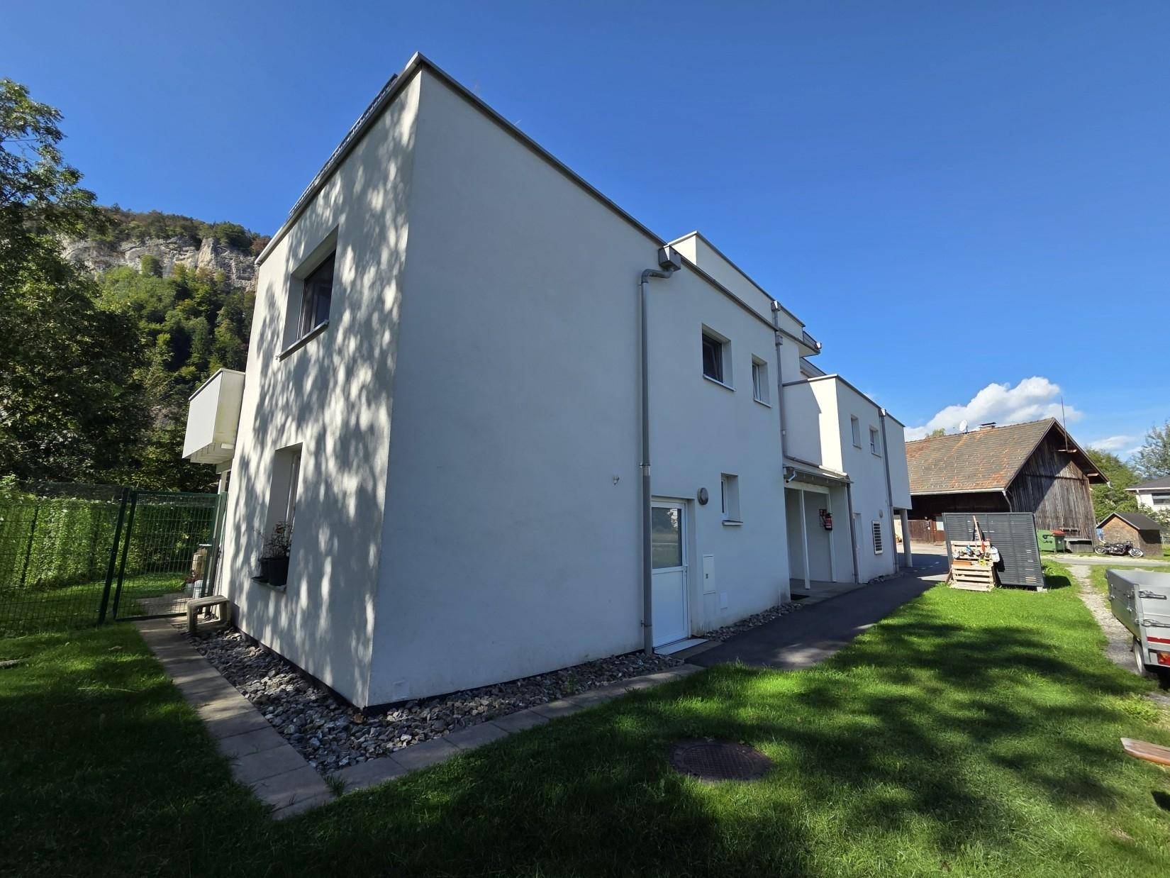 Seitliche Ansicht des modernen Gebäudes mit weißer Fassade, grünem Rasen und traditioneller Scheune im Hintergrund.