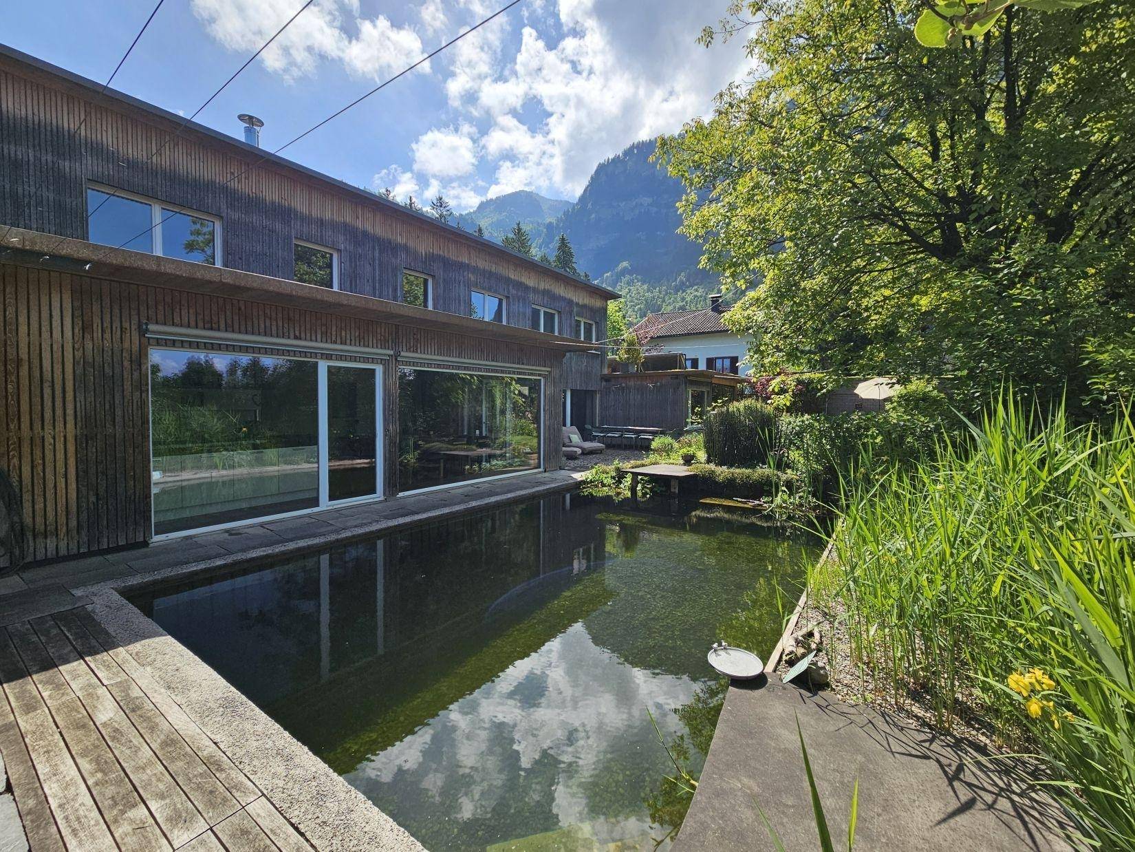 Moderne Holzfassade des Gebäudes mit großen Glasfronten und einem natürlichen Schwimmteich im Vordergrund.