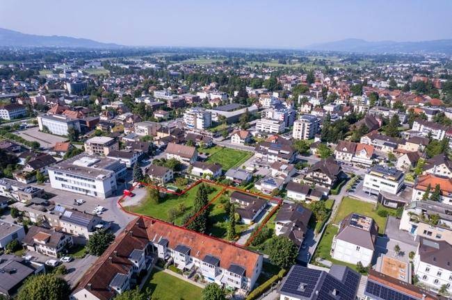 Weitläufige Luftaufnahme einer Stadt mit Bergen im Hintergrund und einem markierten Grundstück.