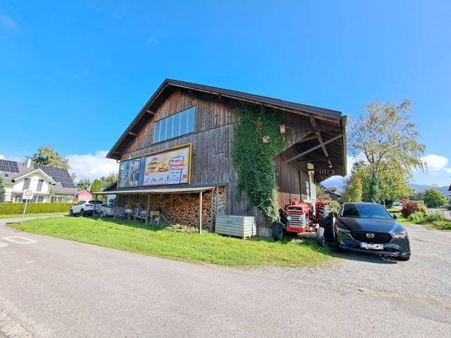 Ansicht des Holzgebäudes mit Werbetafel, Holzlager und geparktem Auto im Freien.