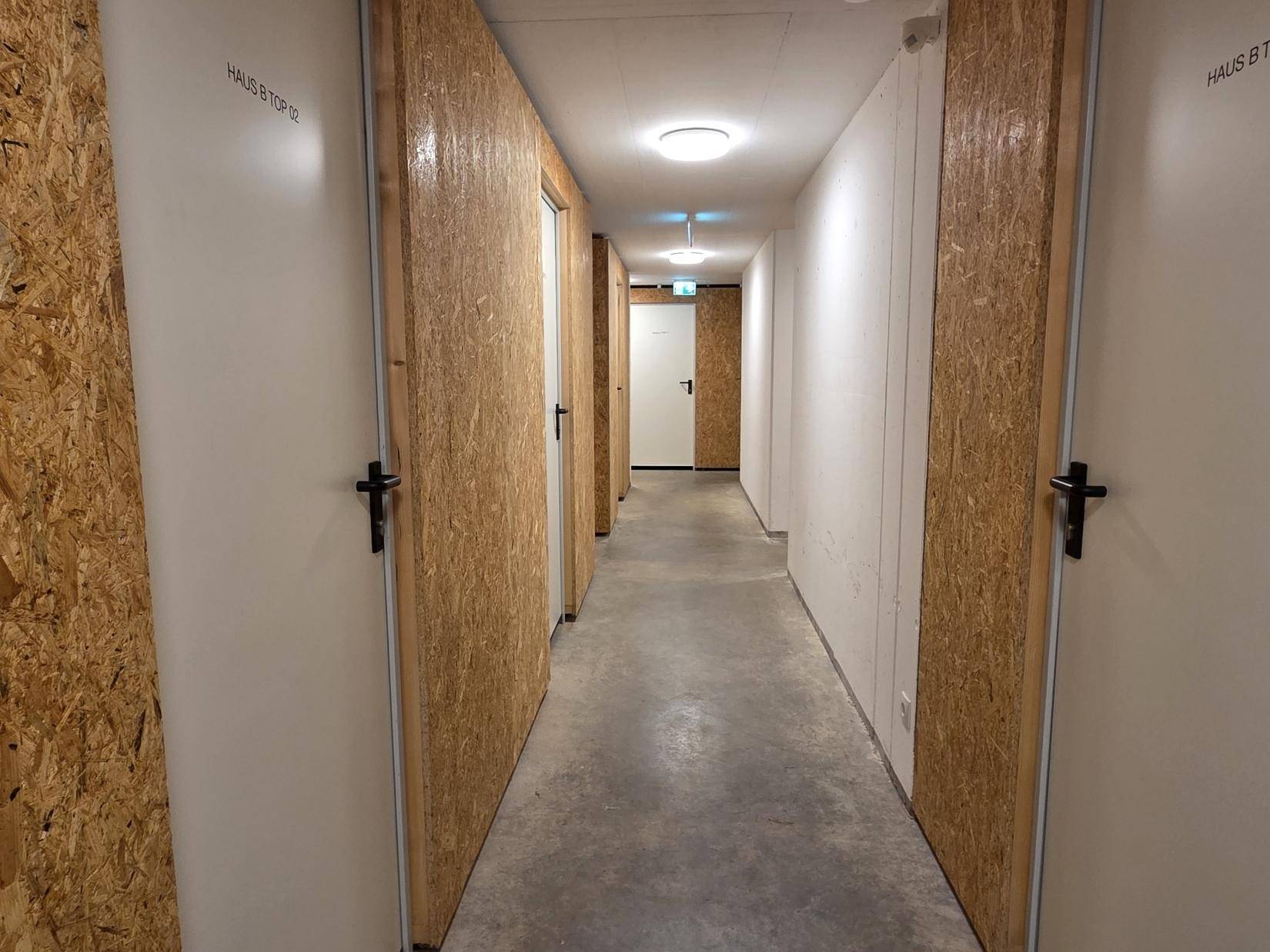 Langer Flur mit individuellen Abstellräumen und Betonboden in einem Mehrfamilienhaus.