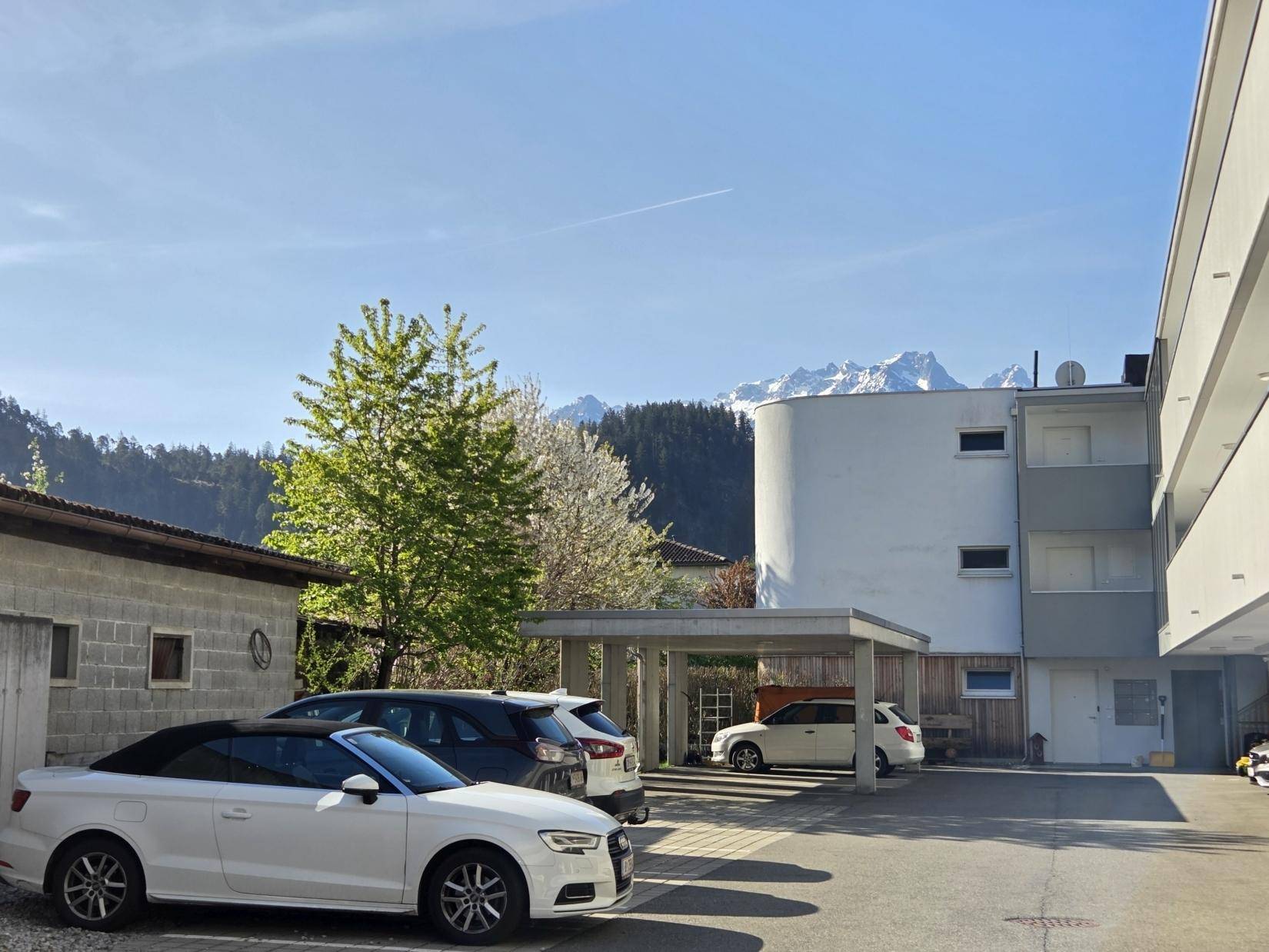 Wohngebäude mit Parkplätzen und Carports, umgeben von Bäumen und Bergen im Hintergrund.