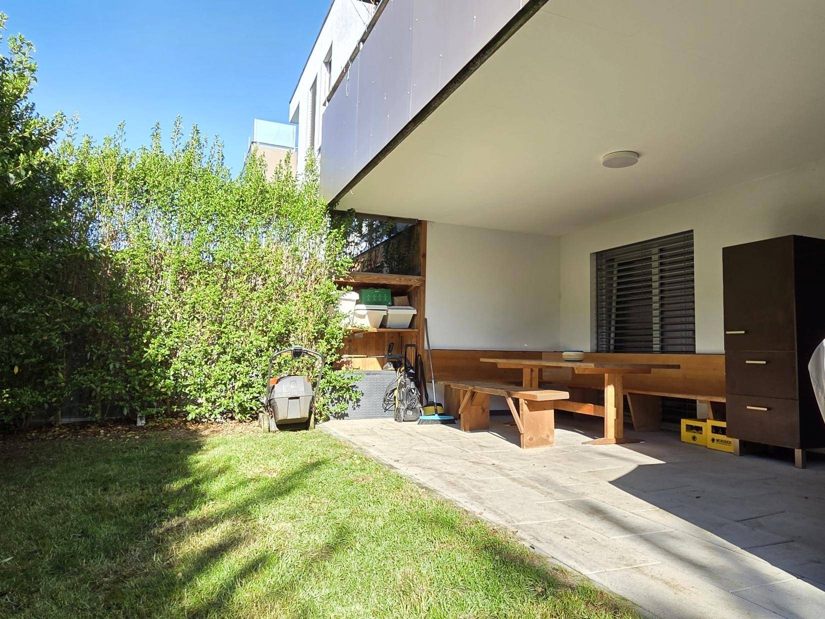 Überdachte Terrasse mit Holztisch und Bänken, angrenzend an einen gepflegten Rasen und Hecke.