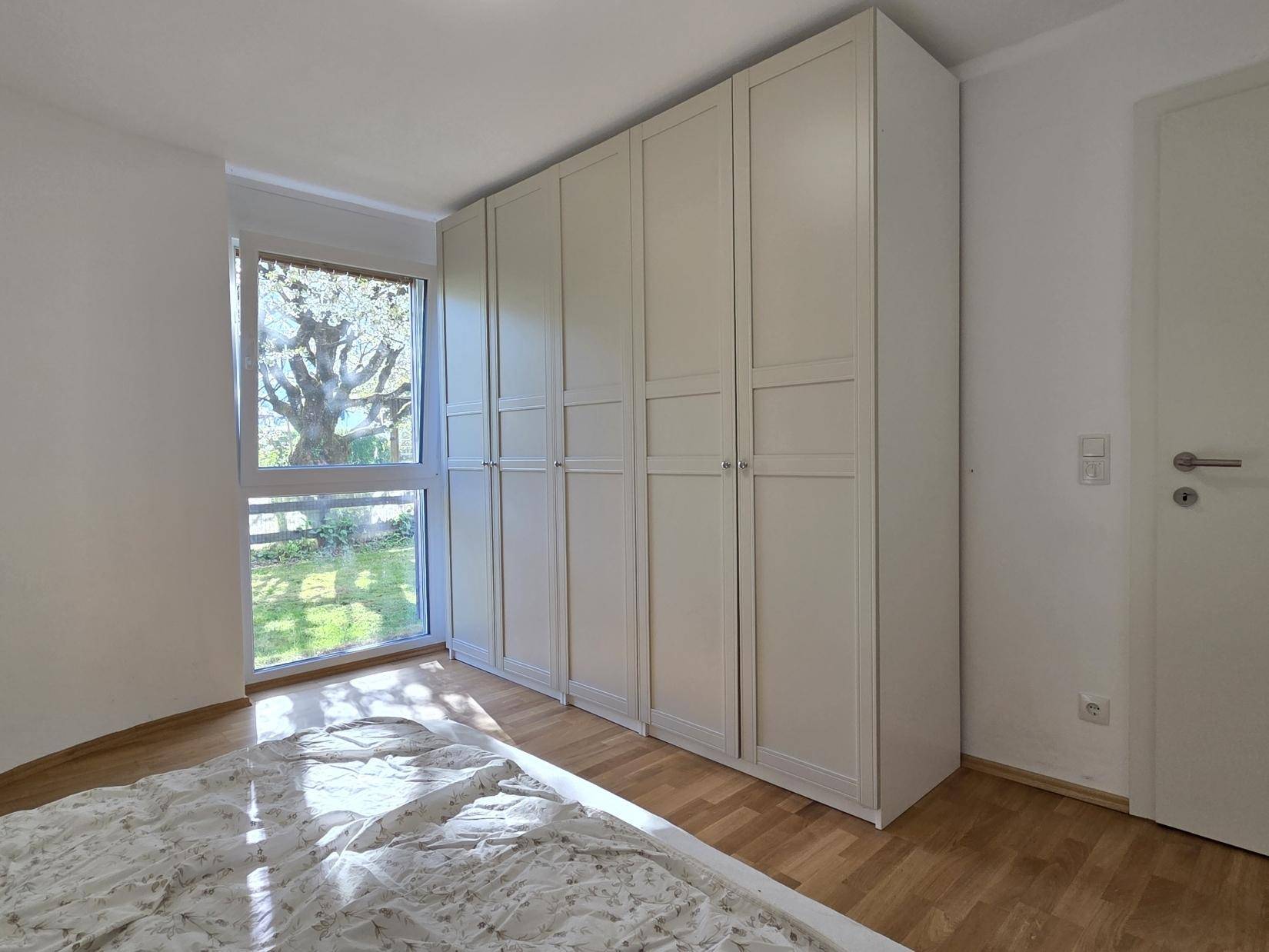 Geräumiges Schlafzimmer mit großem Fenster, weißem Kleiderschrank und Blick ins Grüne.