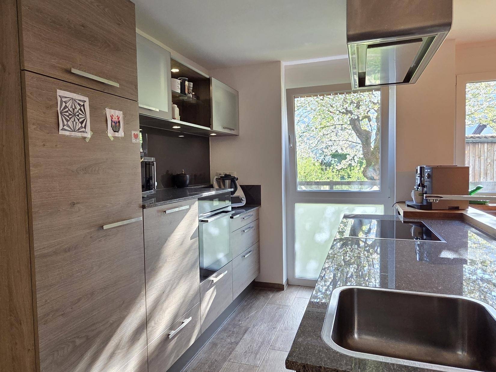 Moderne Küche mit Holzfronten, dunkler Arbeitsplatte und viel Tageslicht durch das Fenster.
