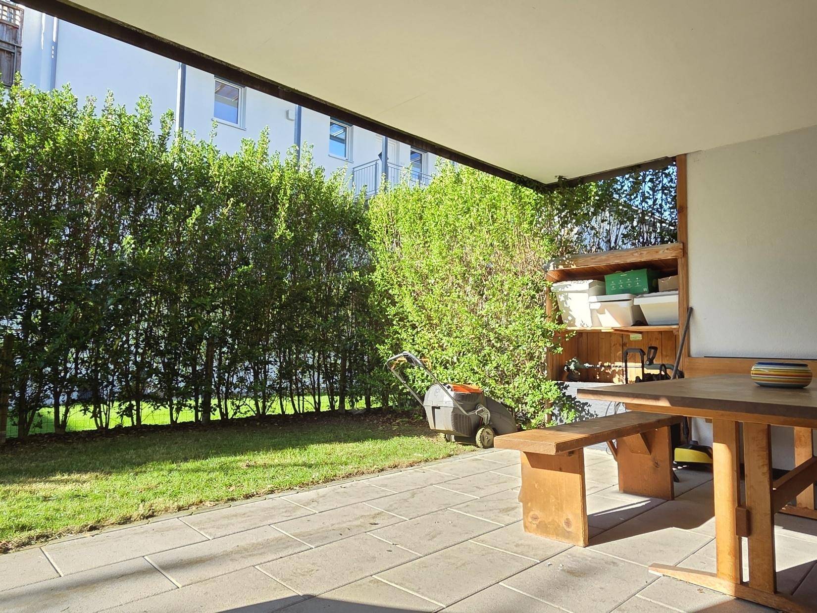 Gemütliche überdachte Terrasse mit Sitzgelegenheiten, umgeben von Grünflächen und einer Hecke.