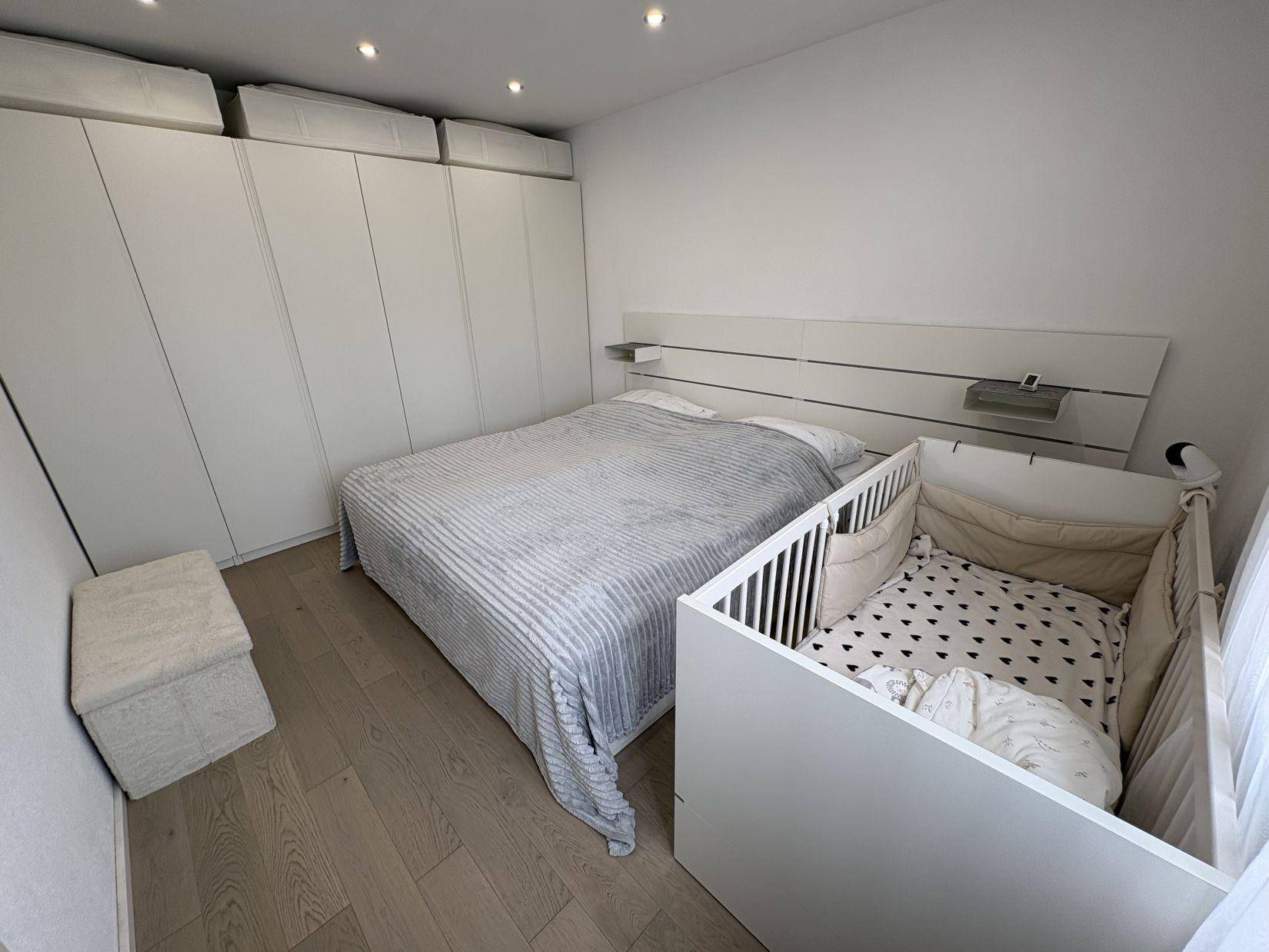 Helles Schlafzimmer mit Doppelbett, Babybett und weißen Einbauschränken.
