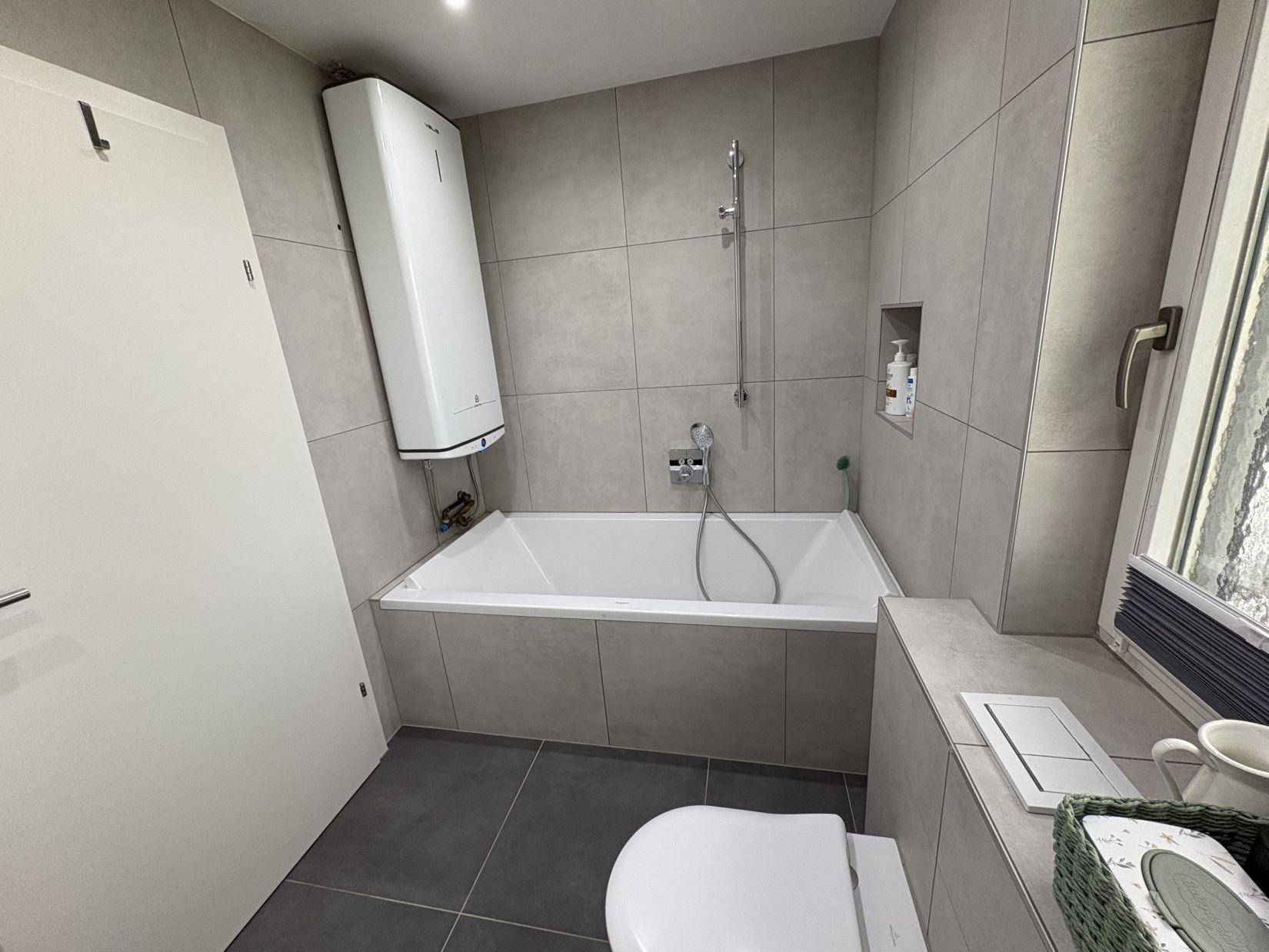 Modernes Badezimmer mit Badewanne, grauen Fliesen und integriertem WC.