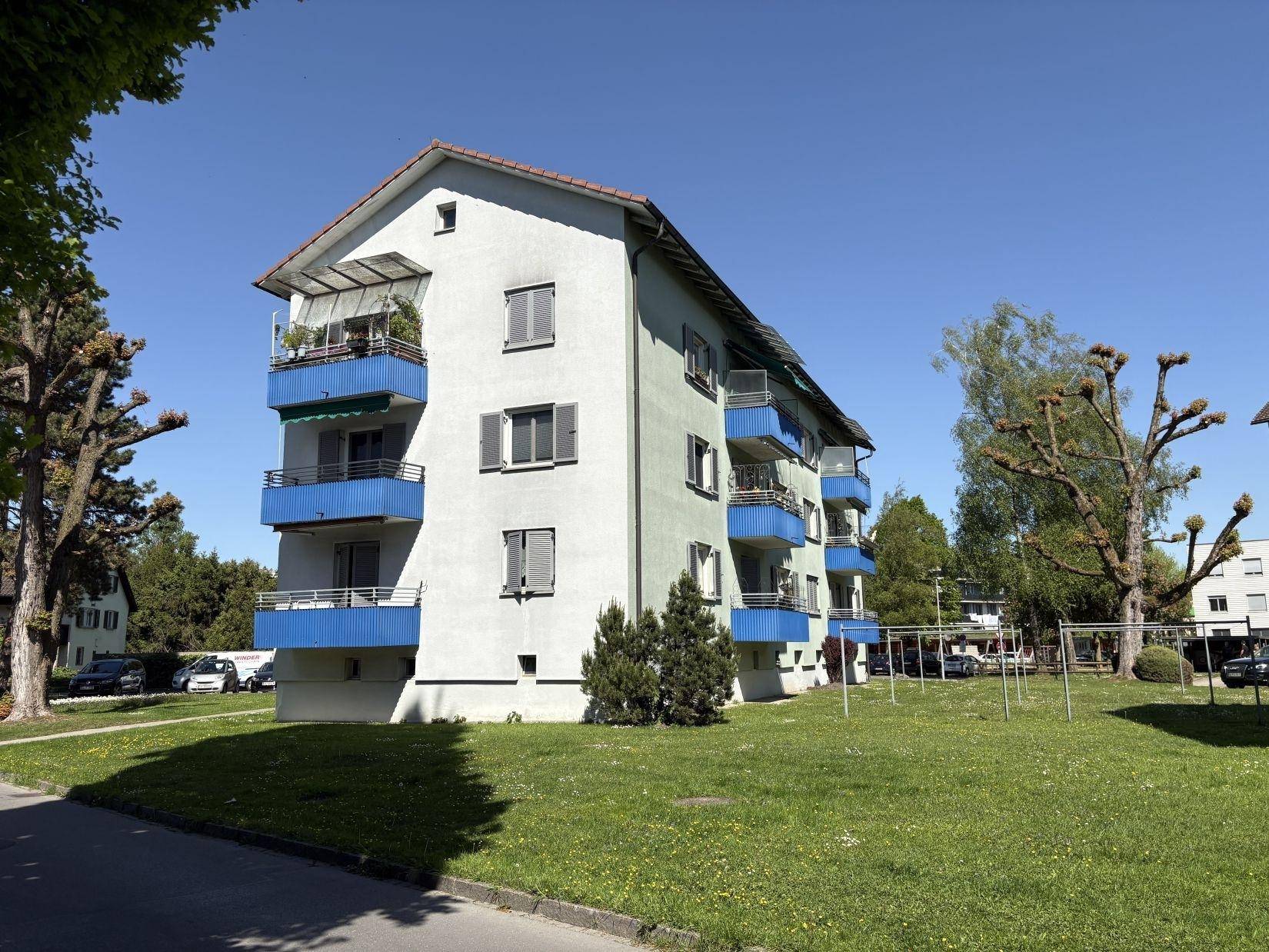 Mehrfamilienhaus mit blauen Balkonen und Spielplatz in grüner Umgebung.