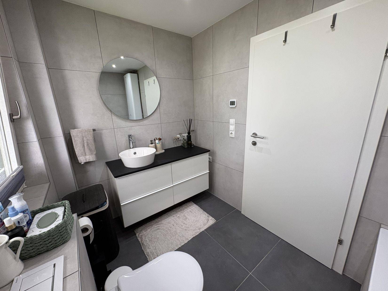 Modernes Badezimmer mit rundem Spiegel, Einzelwaschbecken und grauen Fliesen.