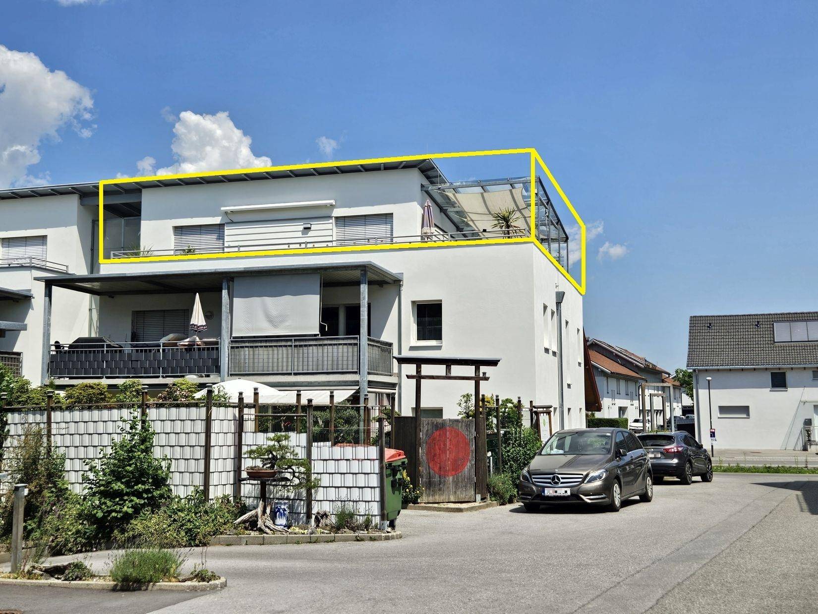 Modernes Wohngebäude mit weißer Fassade und Dachterrasse, umgeben von Grünflächen und Parkplätzen.
