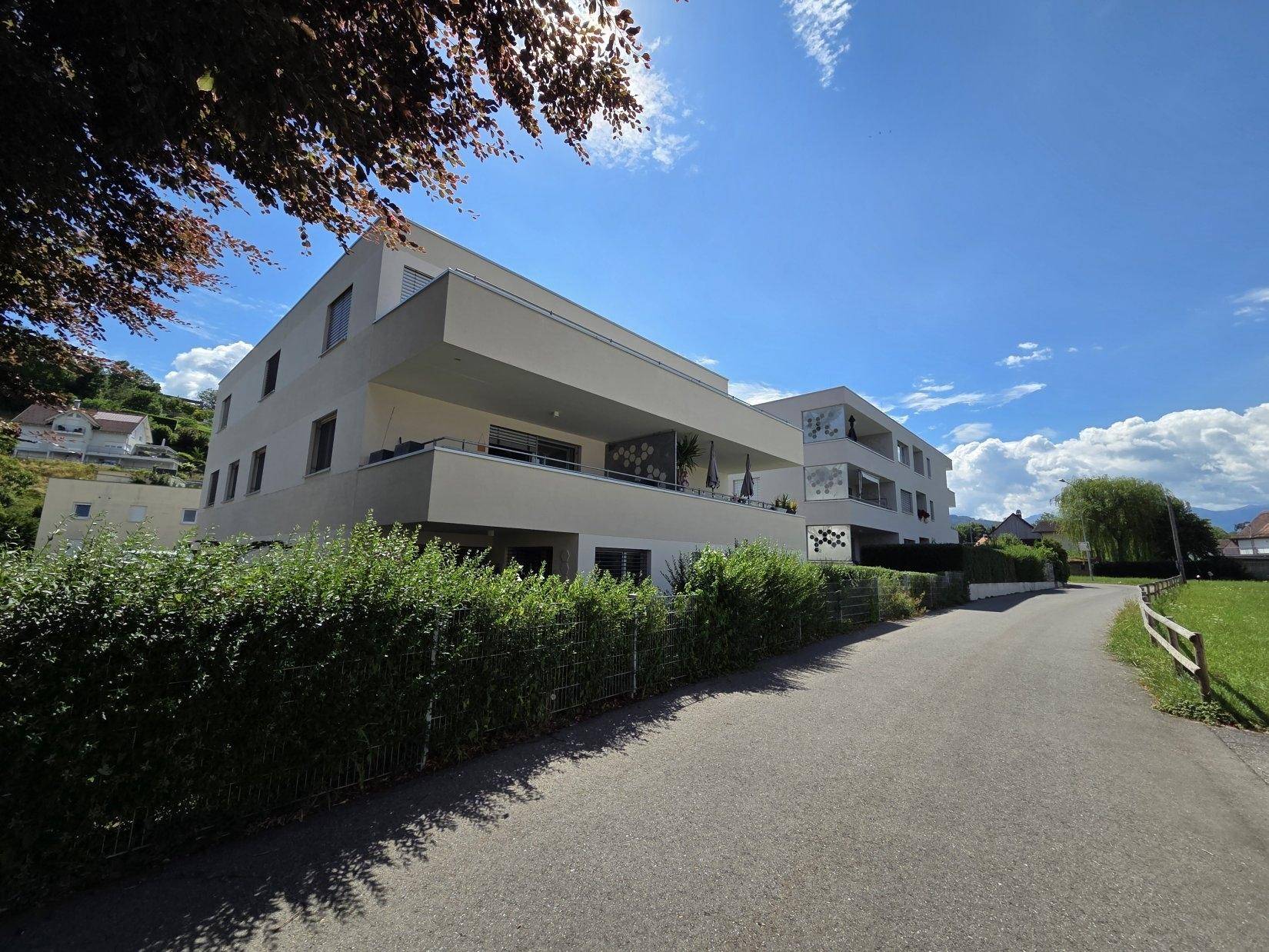 Zwei moderne Wohngebäude mit Balkonen und gepflegten Hecken entlang einer Straße am Hang.